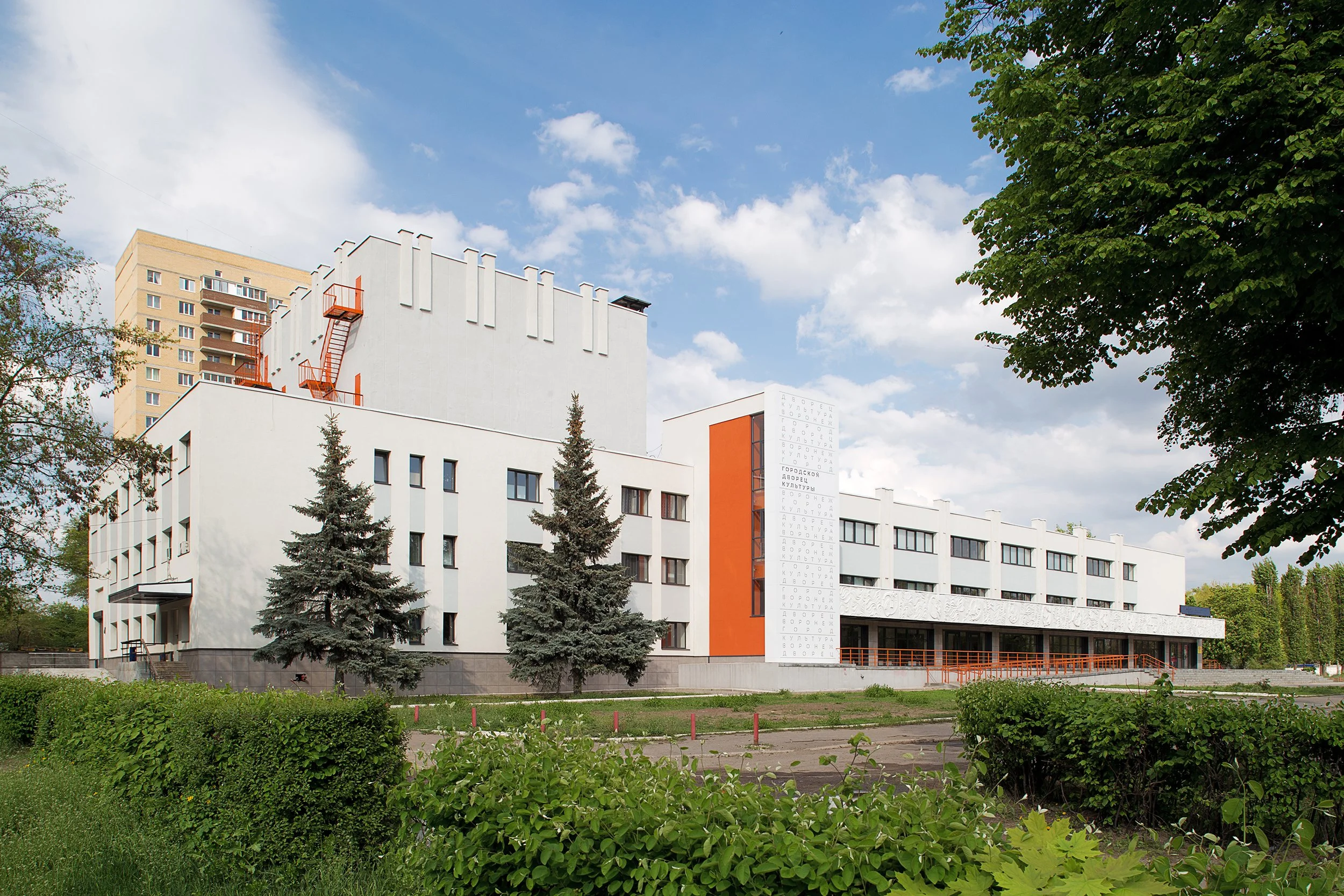 Modern white building with orange accents, surrounded by trees and greenery, under a partly cloudy sky.