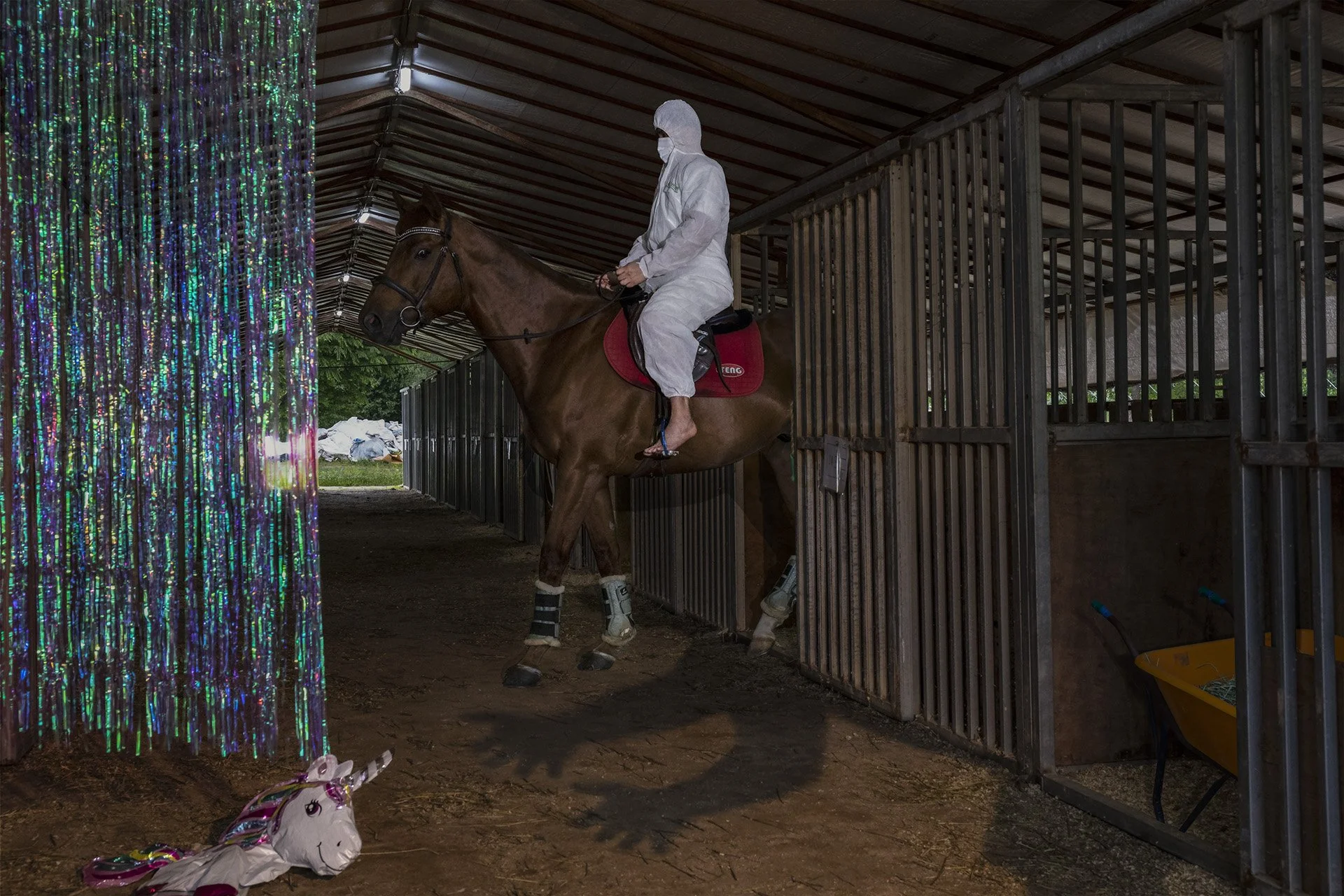 A person in a white protective suit and mask riding a brown horse inside a stable aisle, with a colorful unicorn balloon toy on the ground nearby.