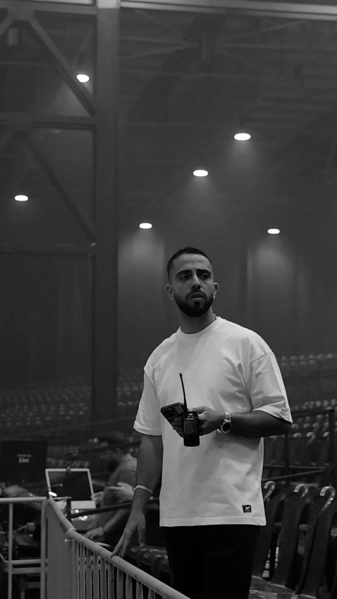 A man with a beard and short hair standing in an indoor setting holding a walkie-talkie. He is wearing a white t-shirt, watch, and bracelets. The background shows an auditorium or conference hall with empty seats, other people using laptops, and some lighting fixtures on the ceiling.