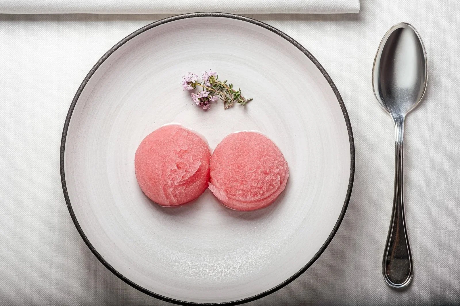 Dos bolas de helado rosado en un plato blanco con un pequeño ramo de flores y una cuchara metálica a la derecha.