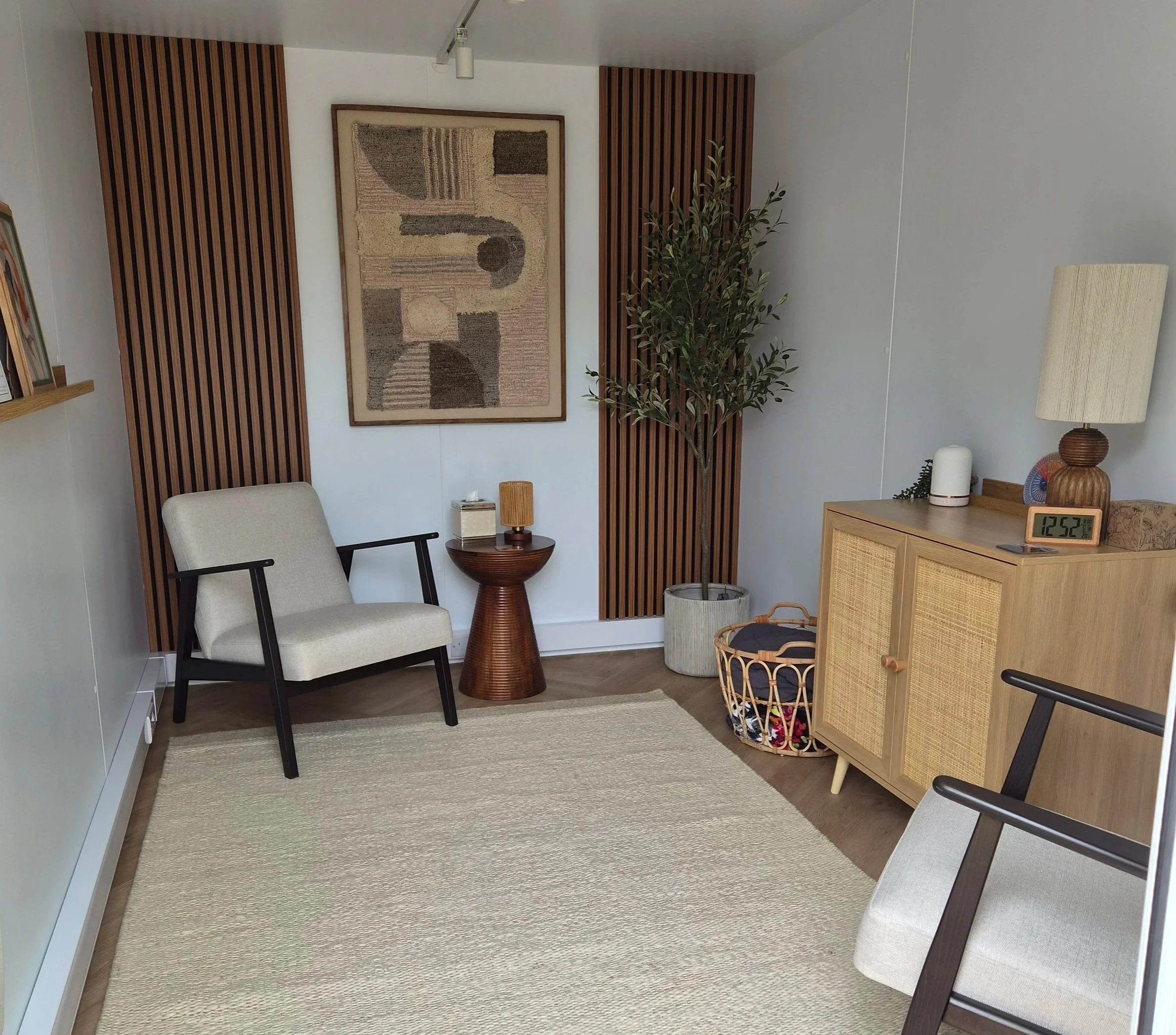 Cozy living room with an accent wall featuring vertical wooden slats, a framed abstract art piece, a potted plant, a beige armchair, a small wooden side table with a lamp, a wicker basket, and a digital clock on a wooden console.