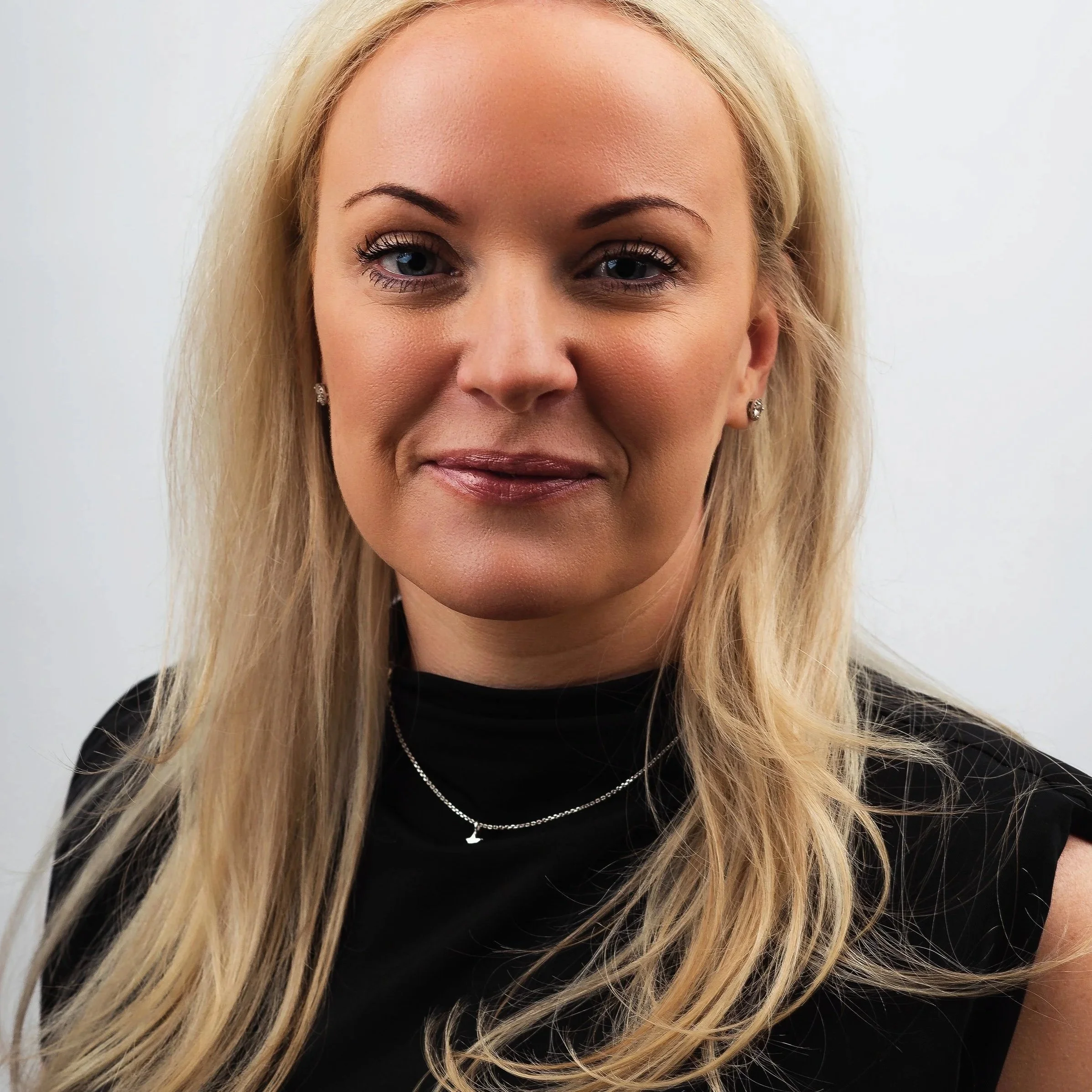 A woman with long blonde hair, wearing a black top, jewelry, and makeup, smiling softly against a plain white background.