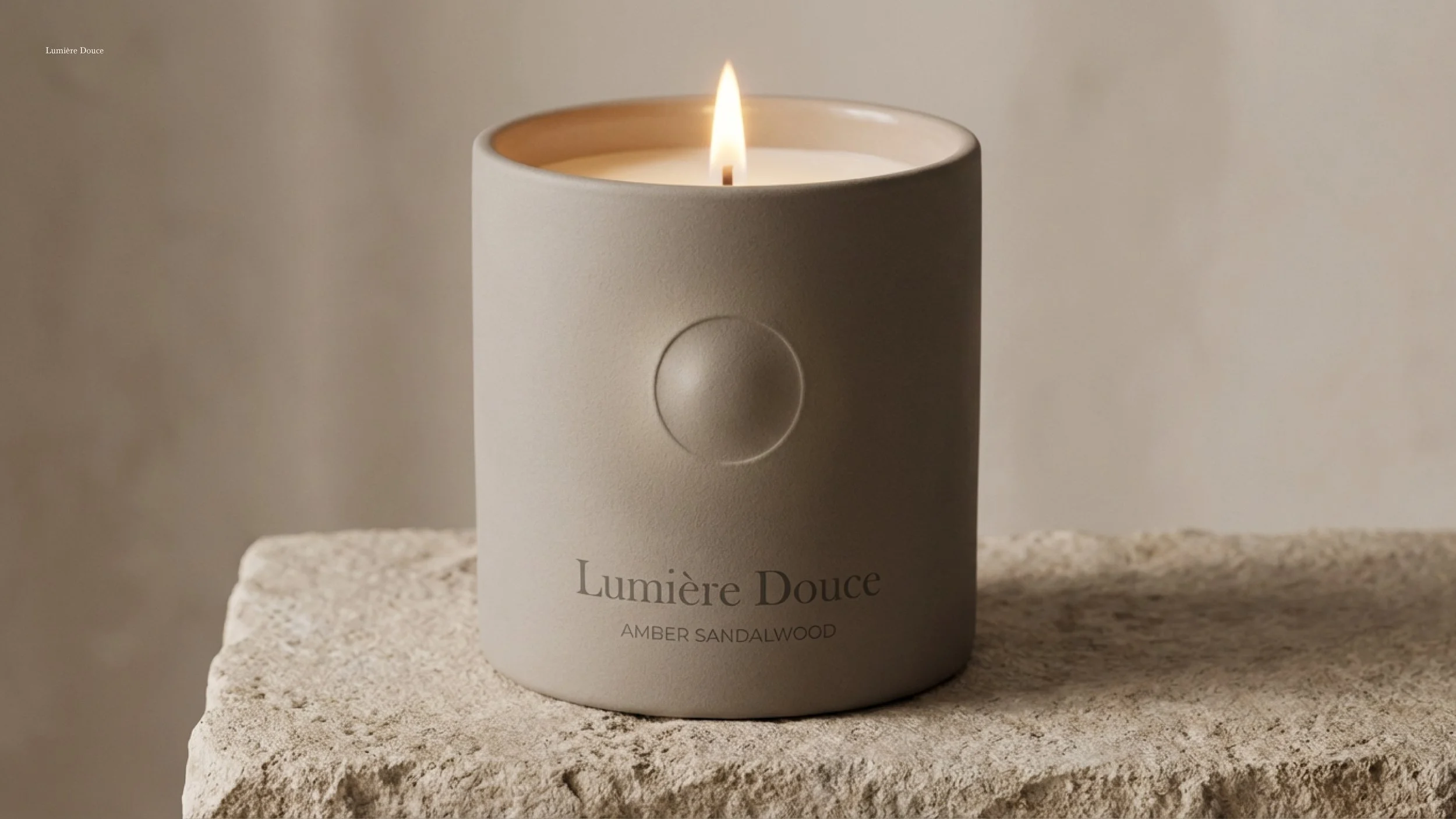 A white candle in a beige container with a lit flame, resting on a rough stone surface. The container has the text "Lumière Douce" and "Amber Sandalwood" printed on it.