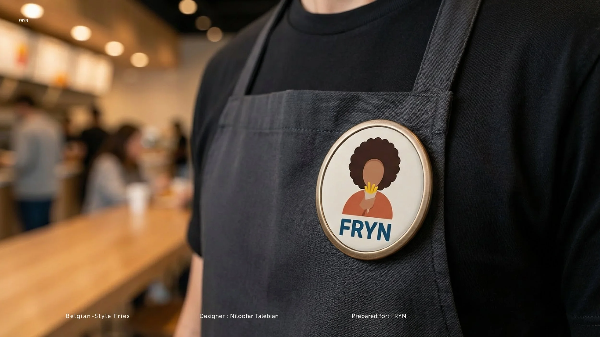 Close-up of a person wearing a black apron with a badge featuring a cartoon woman with curly hair and the word 'FRYN' in blue letters. The background shows a blurred café or restaurant setting with other people.