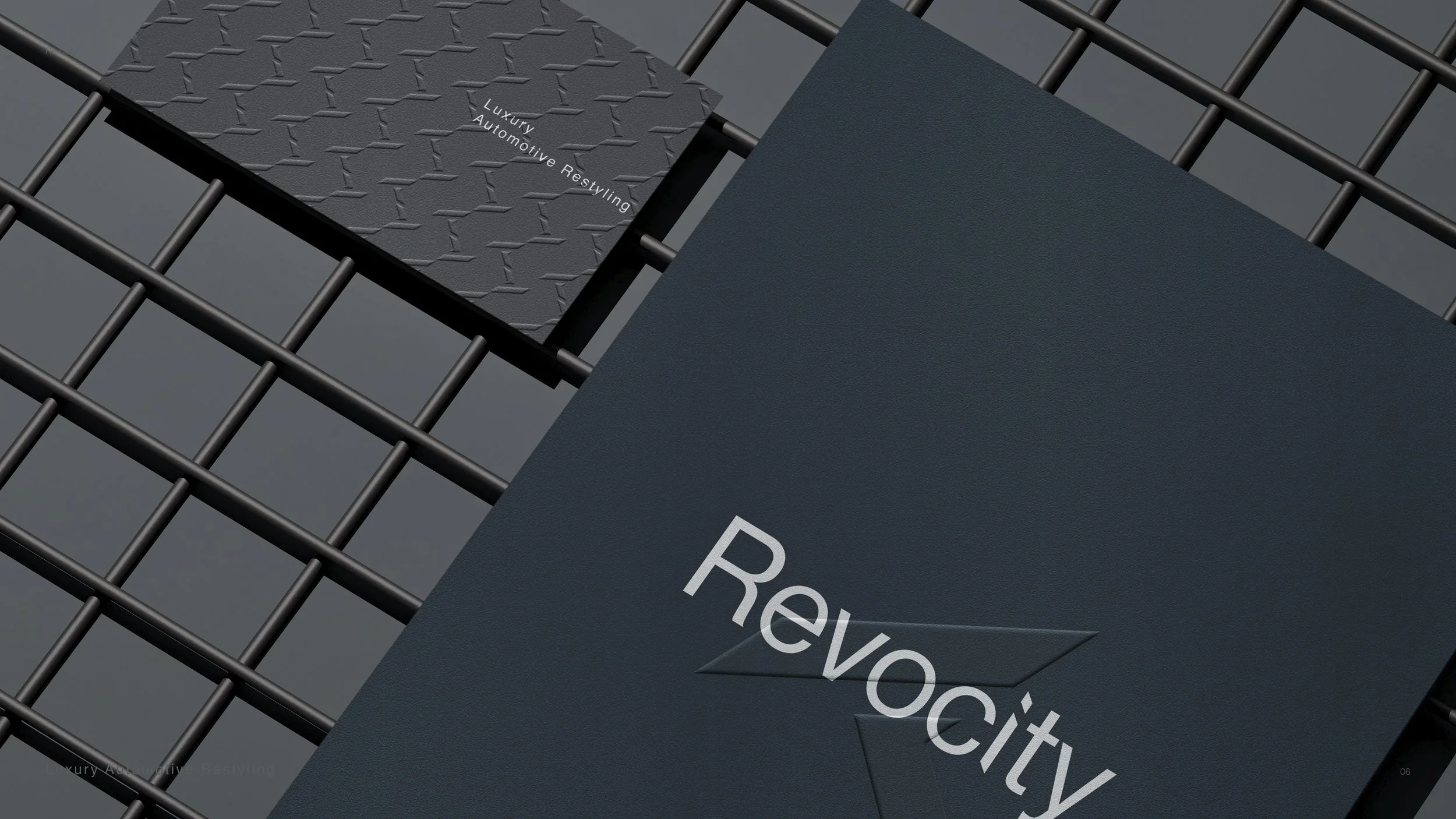 A dark blue booklet with the word 'RevoCity' embossed on the cover, lying on a black metal grid surface. Behind it, a smaller black booklet with the text 'Luxury Automotive Restyling' embossed and a textured pattern.