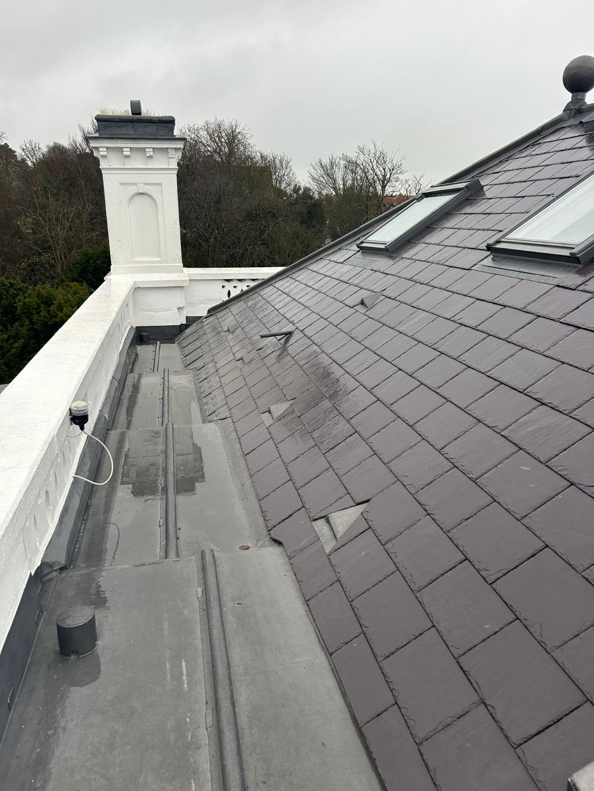 View of a rooftop with purple shingles, skylights, and a white chimney on a cloudy day.