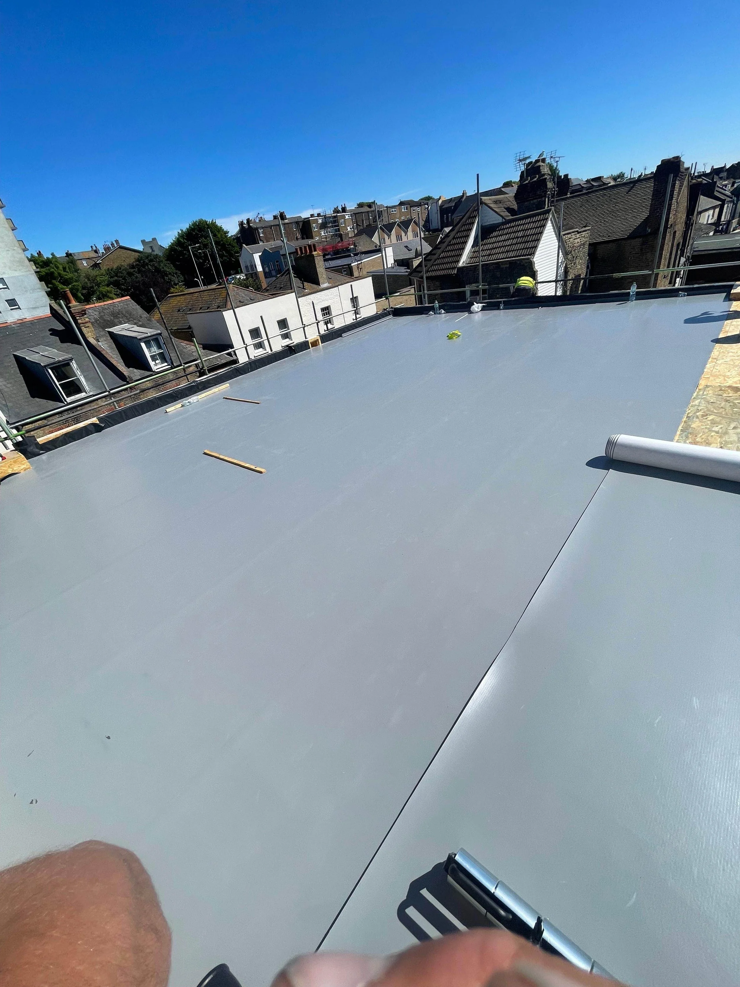 Flat rooftop with a waterproof membrane, some scattered tools, and a roller, surrounded by a safety railing, with neighboring buildings and a clear blue sky in the background.