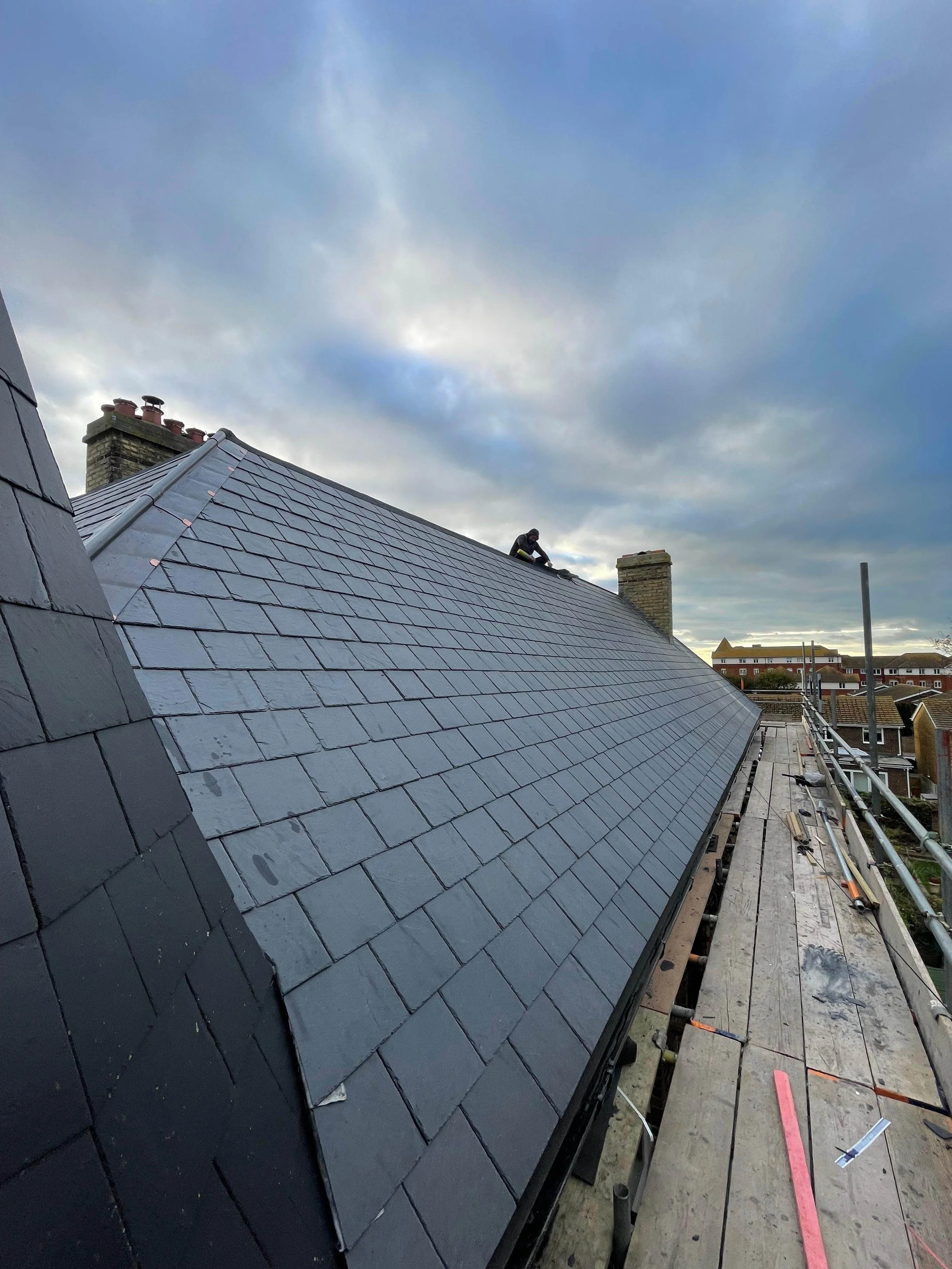 A worker installing or inspecting slate roof tiles on a rooftop with scaffolding, overlooking a neighborhood with residential buildings under a cloudy sky.