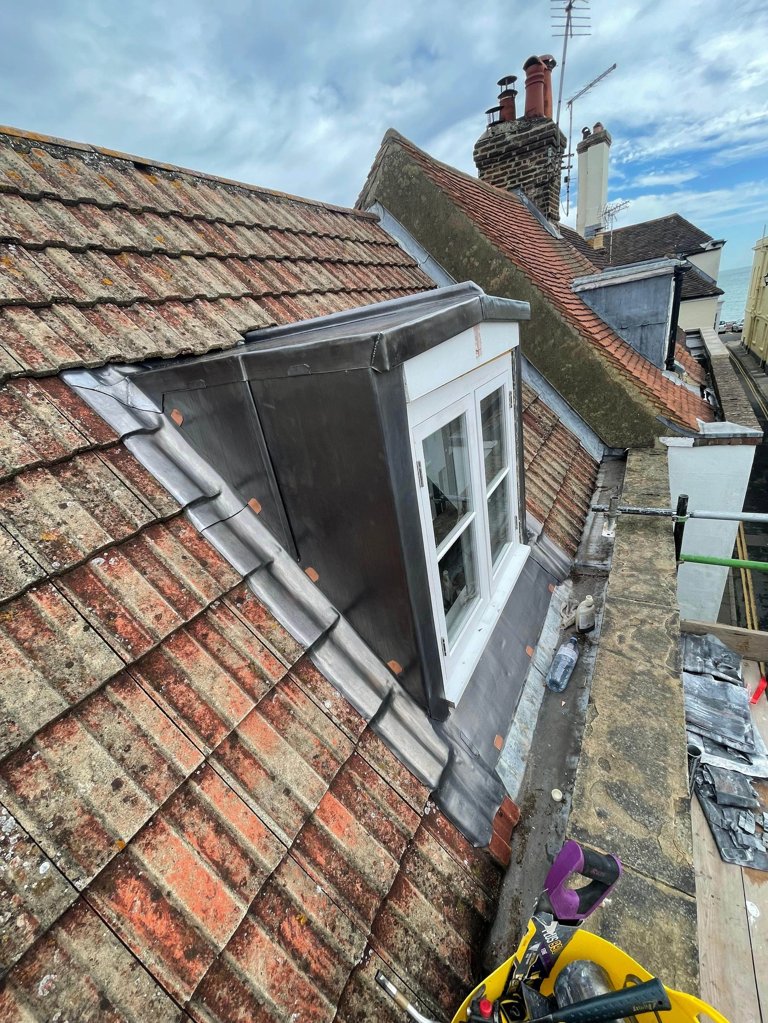 View of a roof with a new dormer window installation, showing old weathered tiles and a stormy sky.