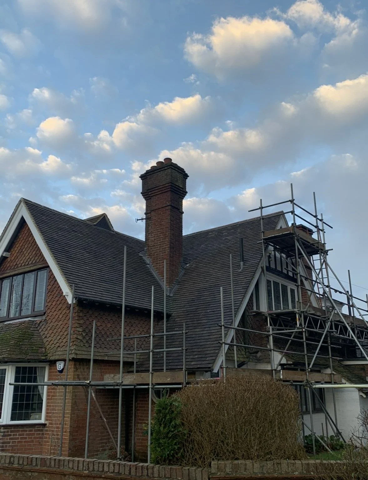 House with a steeply pitched roof, brick chimney, scaffolding around the roof, cloudy sky in the background.