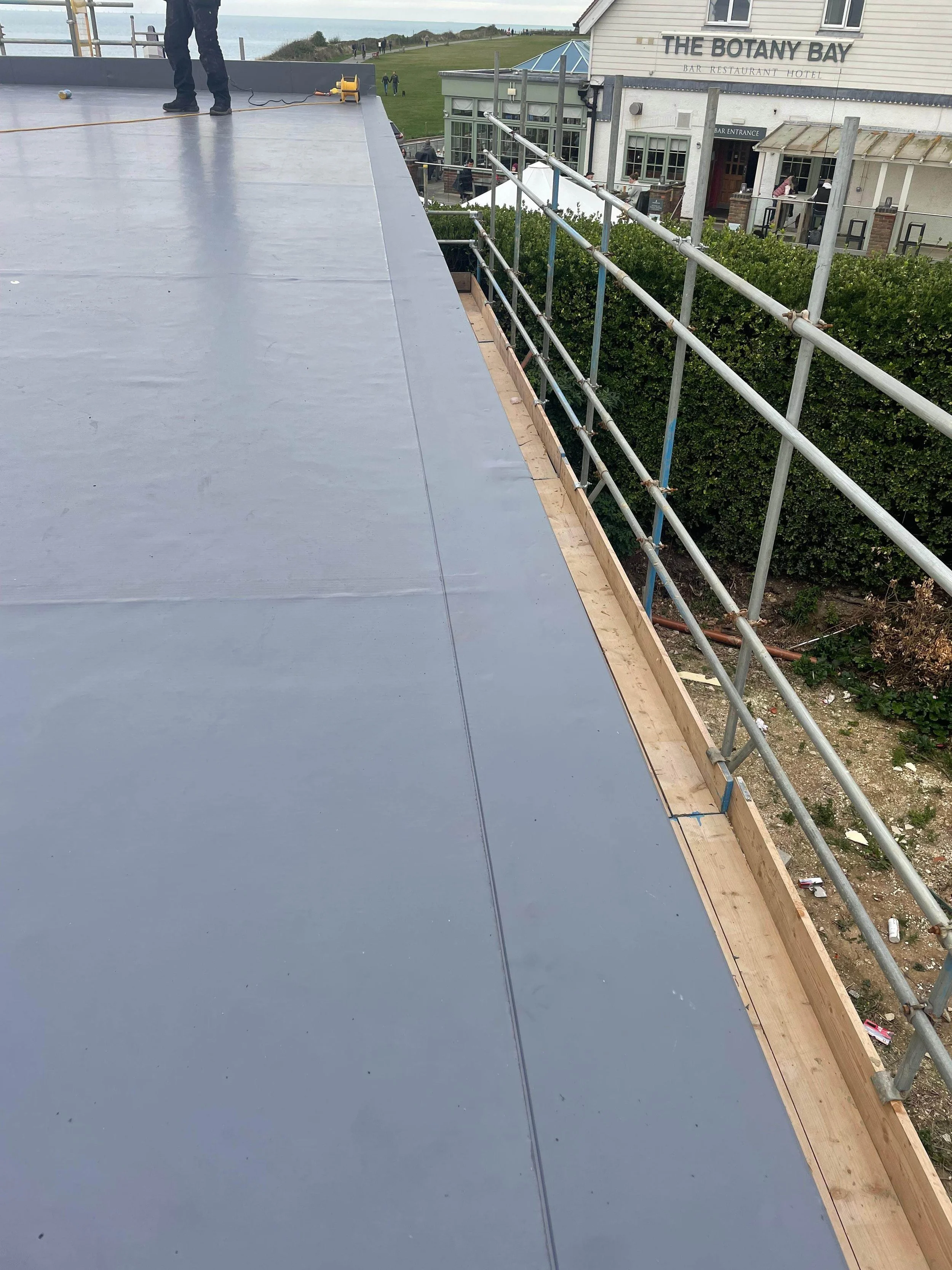 Newly installed flat roofing with a gray surface, surrounded by scaffolding at the edge, on a building near a restaurant called 'The Botany Bay' with outdoor seating, grassy area, and ocean in the background.