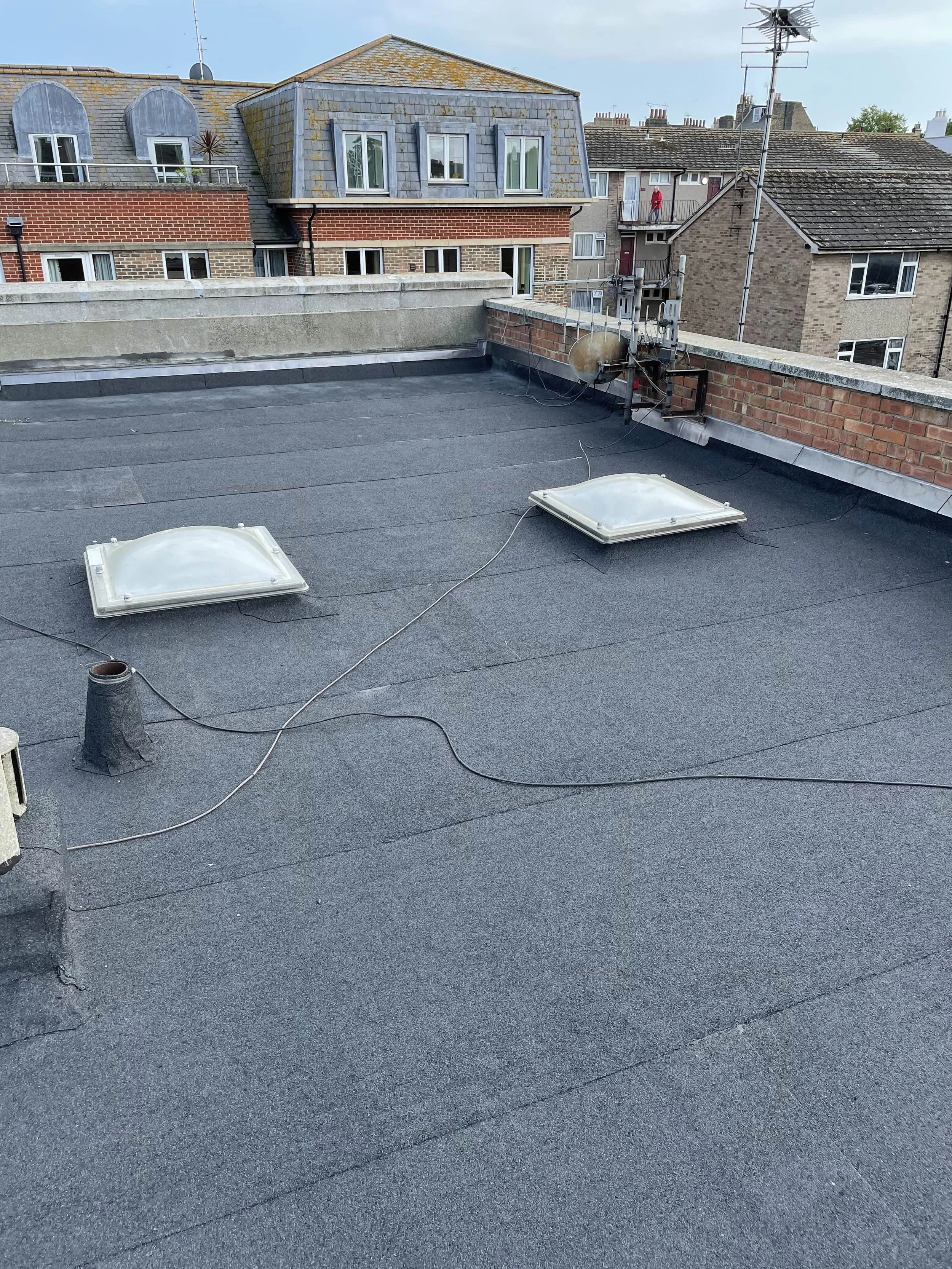 View of a flat rooftop with two skylights, an exhaust pipe, and several electrical wires, surrounded by other residential buildings in the background.