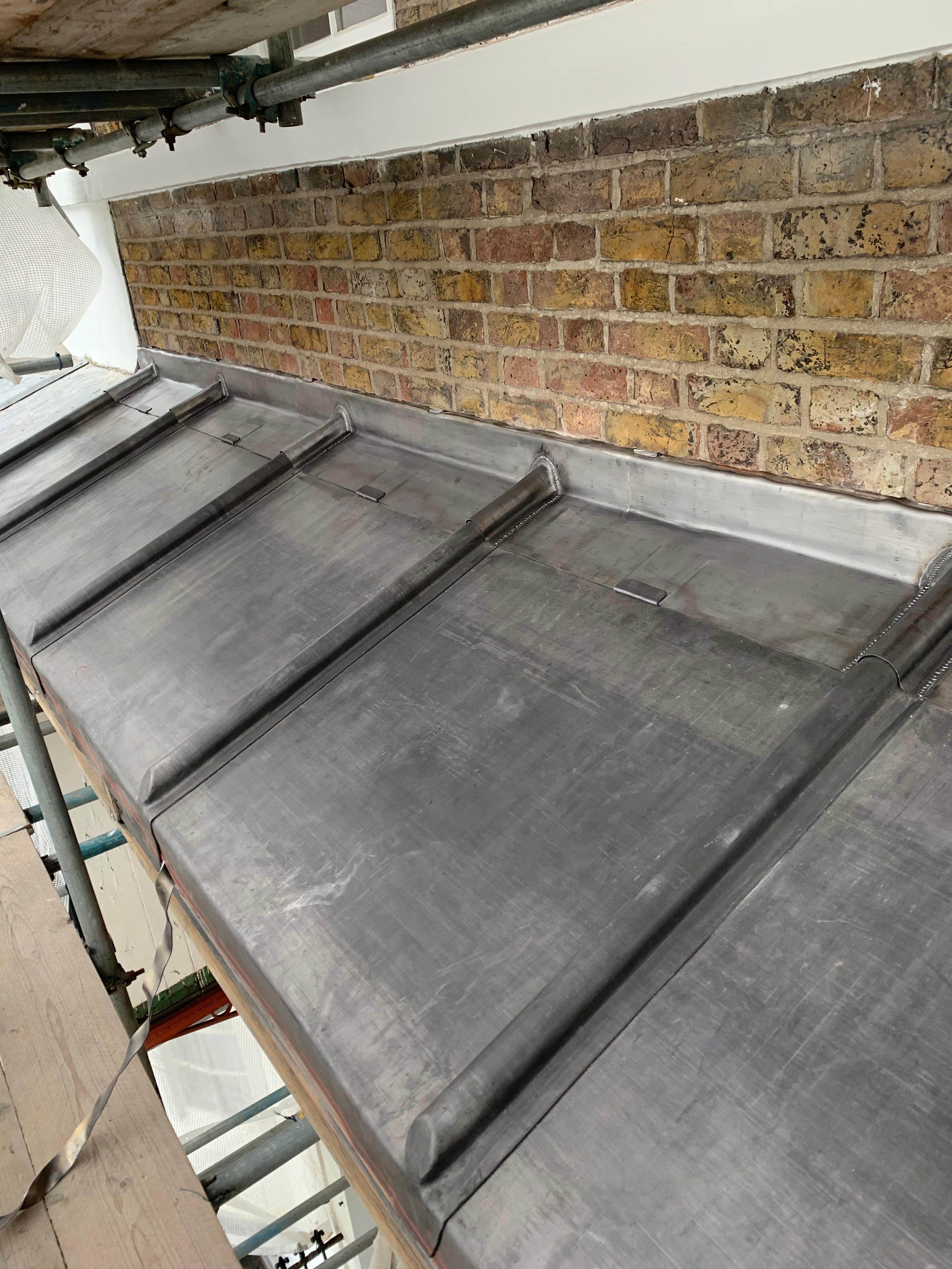 Metal roofing sheets installed on a building with a brick wall in the background, scaffolding around the roof, and construction materials nearby.