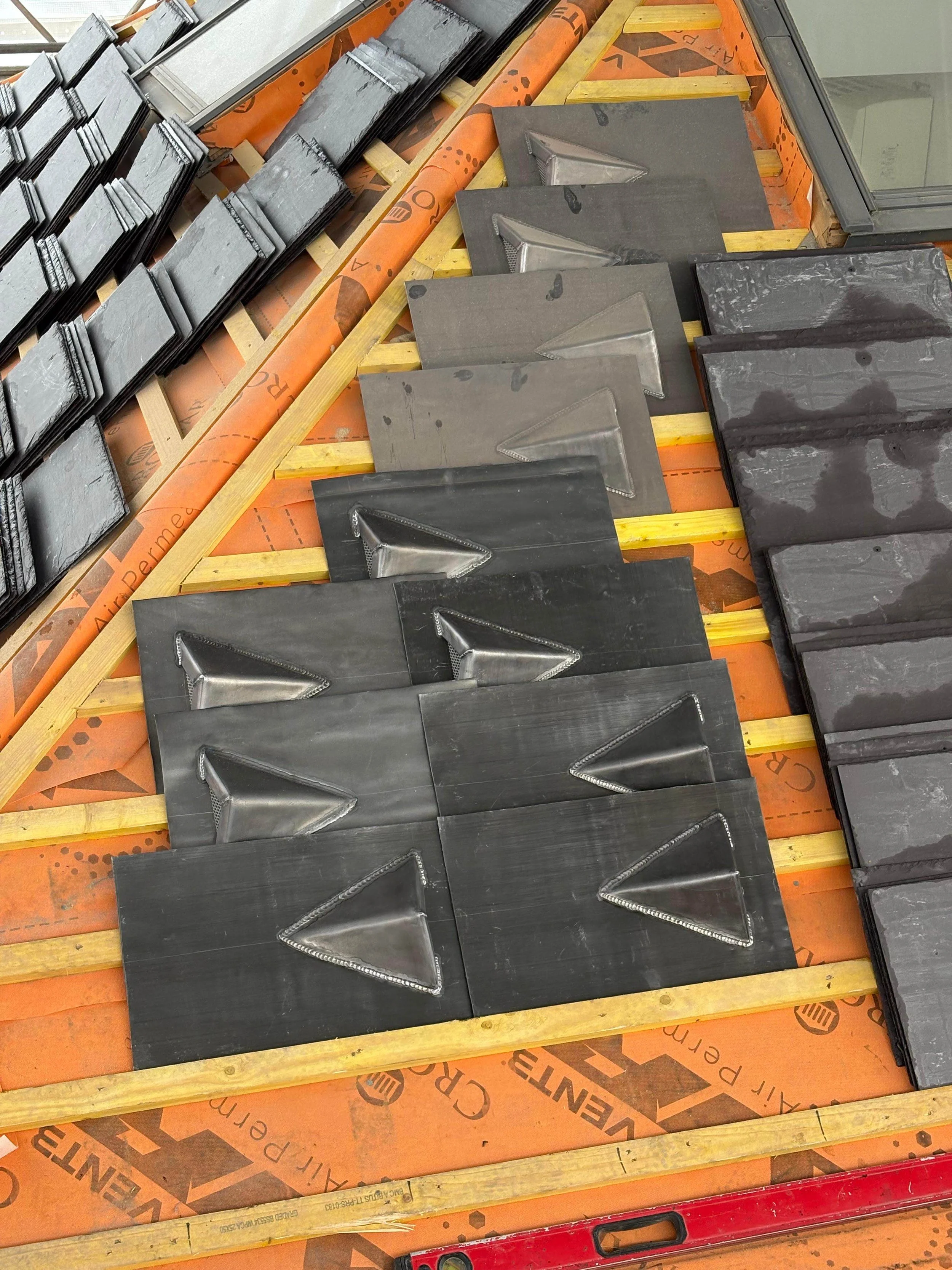 Several black slate tiles with metal pyramid-shaped accents are installed on a roof under construction, supported by yellow wooden battens and orange protective sheathing.