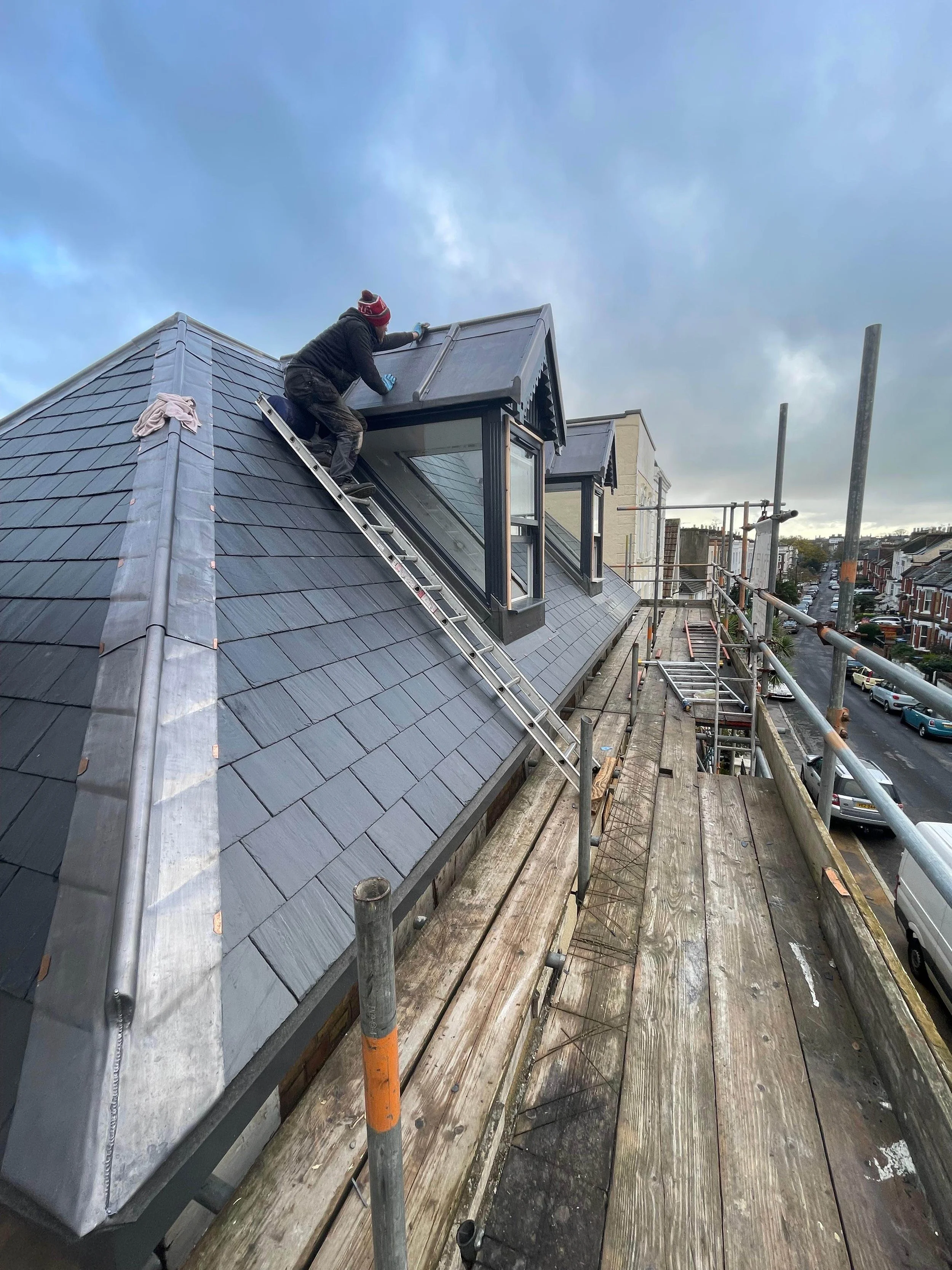 A worker standing on scaffolding, repairing a sloped tiled roof with dormer windows during daytime.