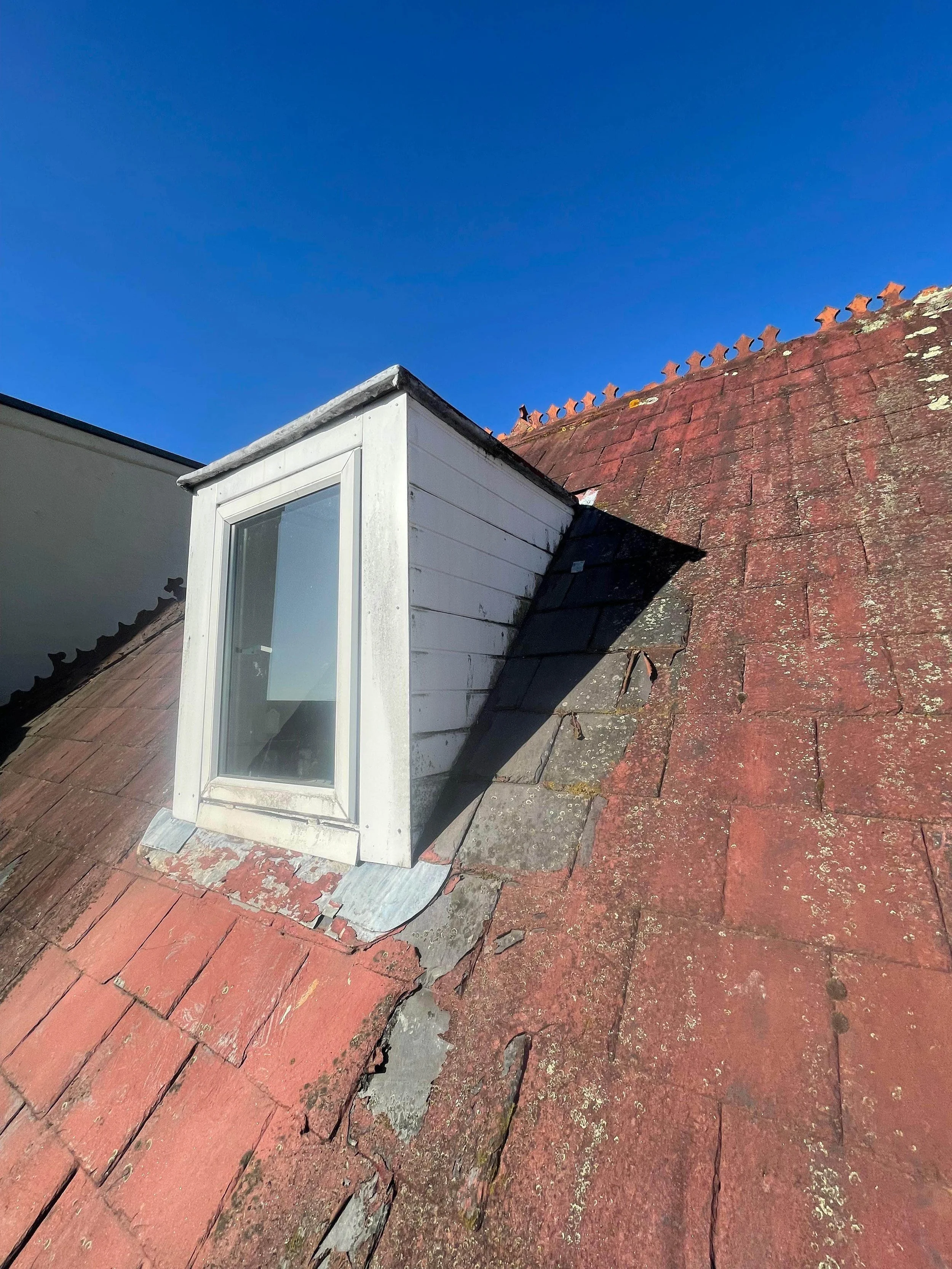 Close-up of a roof with a dormer window, showing damaged and weathered red tiles and shingles, with clear blue sky in the background.