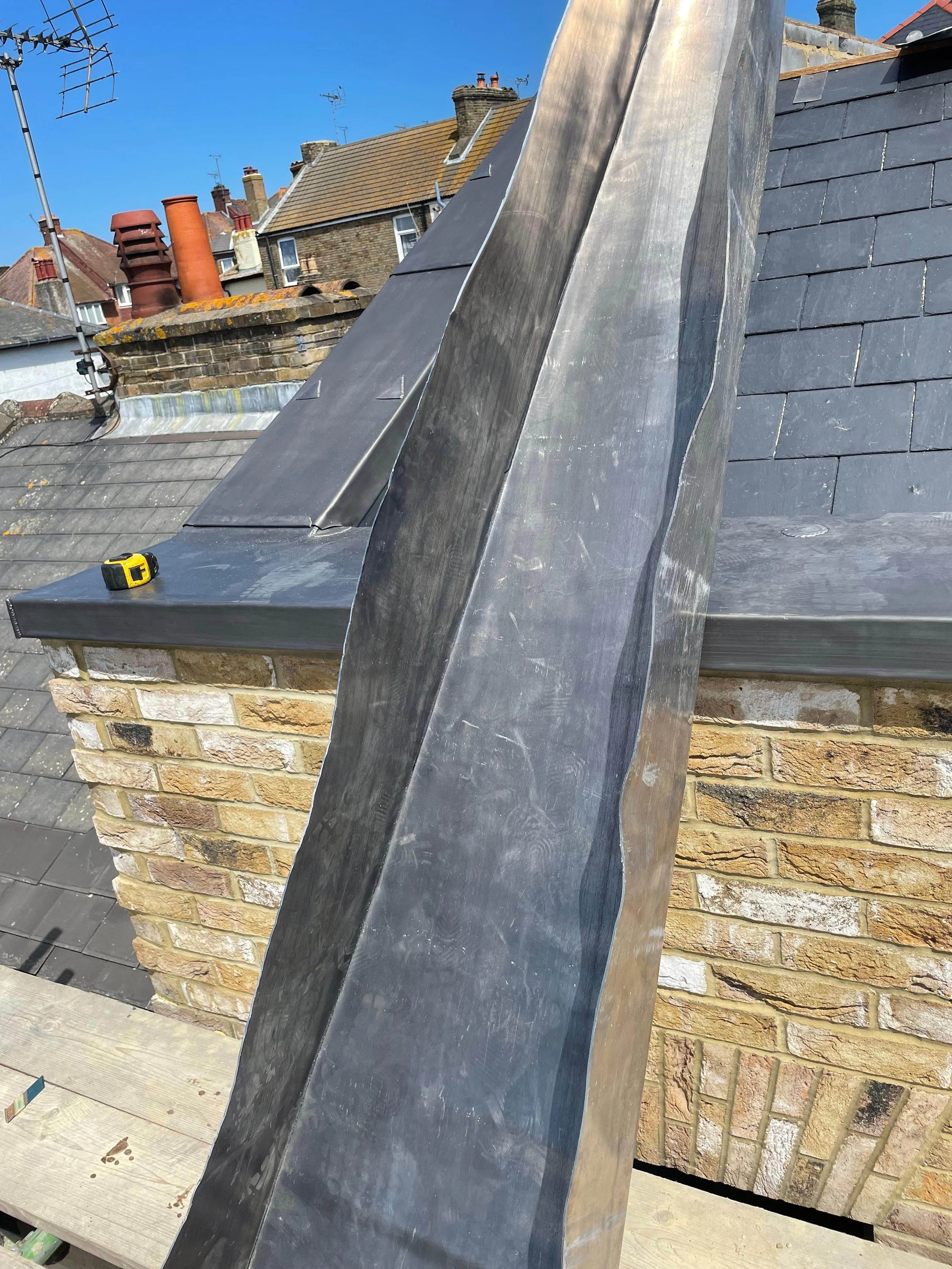 Close-up of a chimney with a new metal flashing installation on a rooftop with neighboring rooftops in the background.