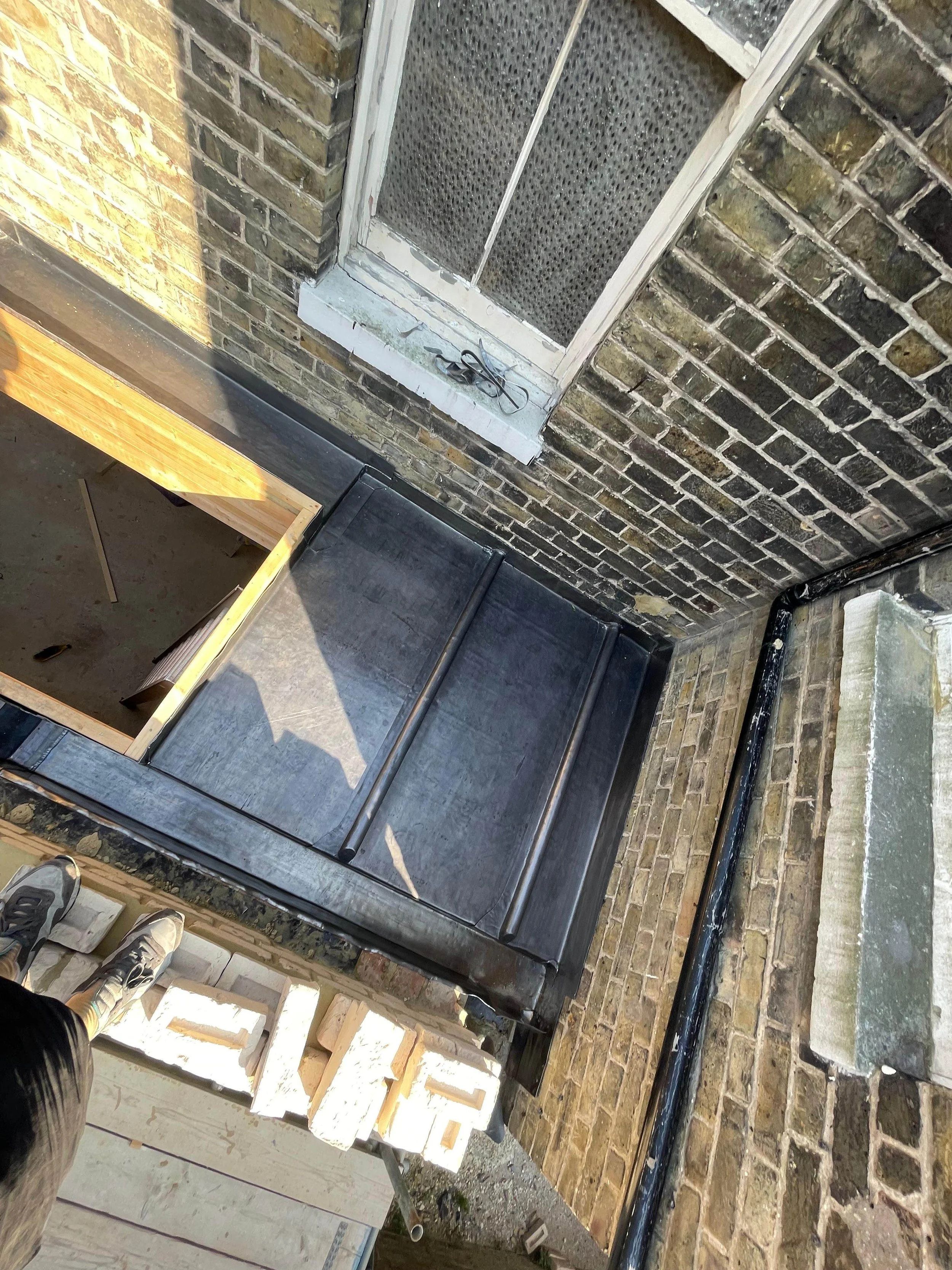 View of a balcony or small outdoor space with brick walls, a window with metal screen, a black flat roof surface, and some construction materials like bricks and wood.
