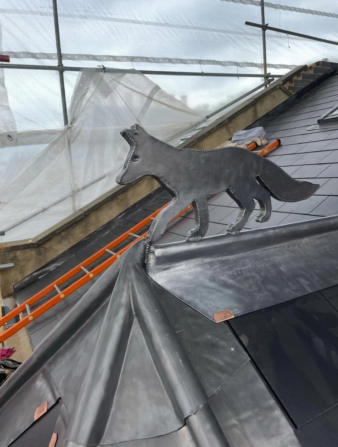 Metal wolf sculpture on a roof made of black metal shingles, with construction scaffolding and a white plastic covering in the background.