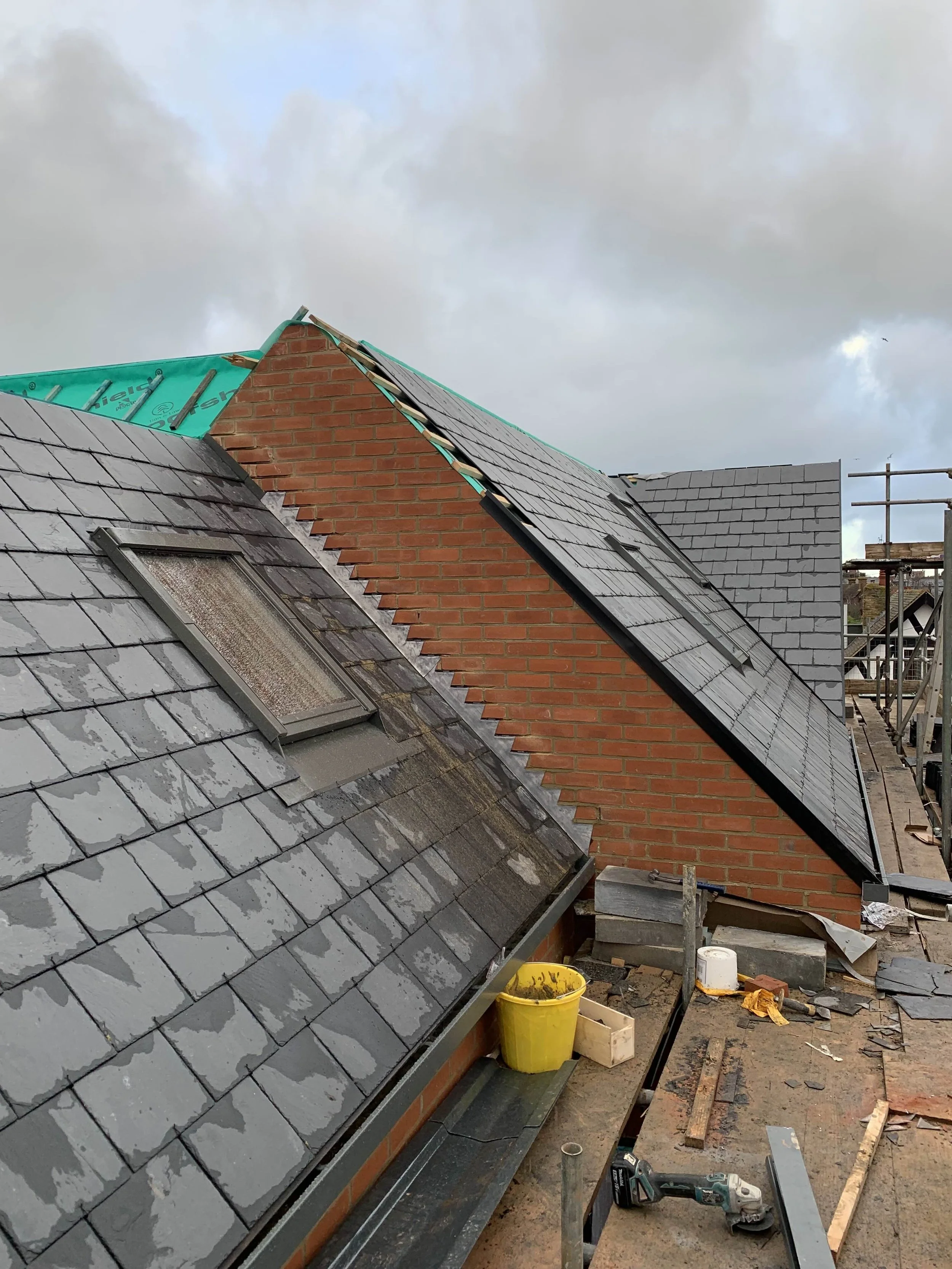 Roof construction with partially installed slate tiles and a skylight, wooden scaffolding and construction tools in the foreground, cloudy sky overhead.