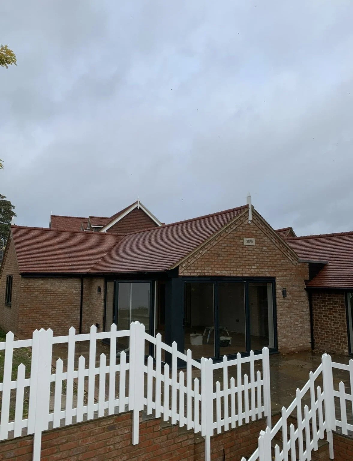 A brick house with a red tile roof, large sliding glass doors, and a white picket fence, under a cloudy sky.