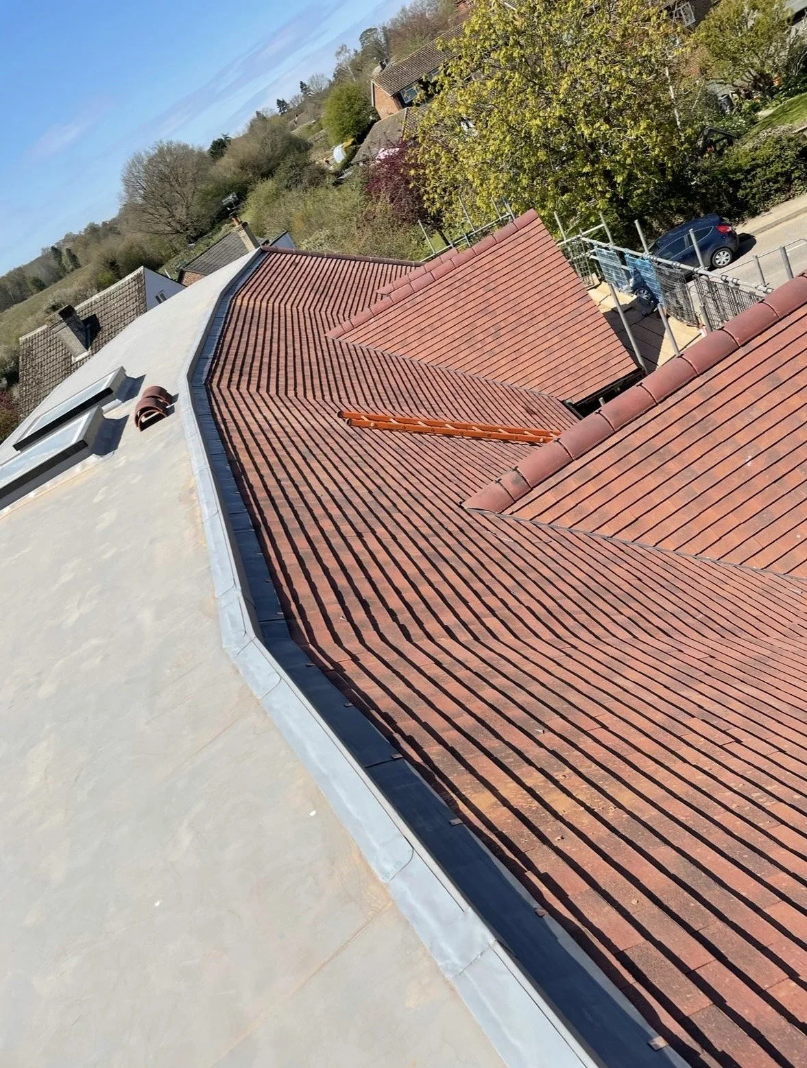 A photograph of a roof with red tiles, seen from an elevated angle on a sunny day. Surrounding landscape includes trees and other houses.