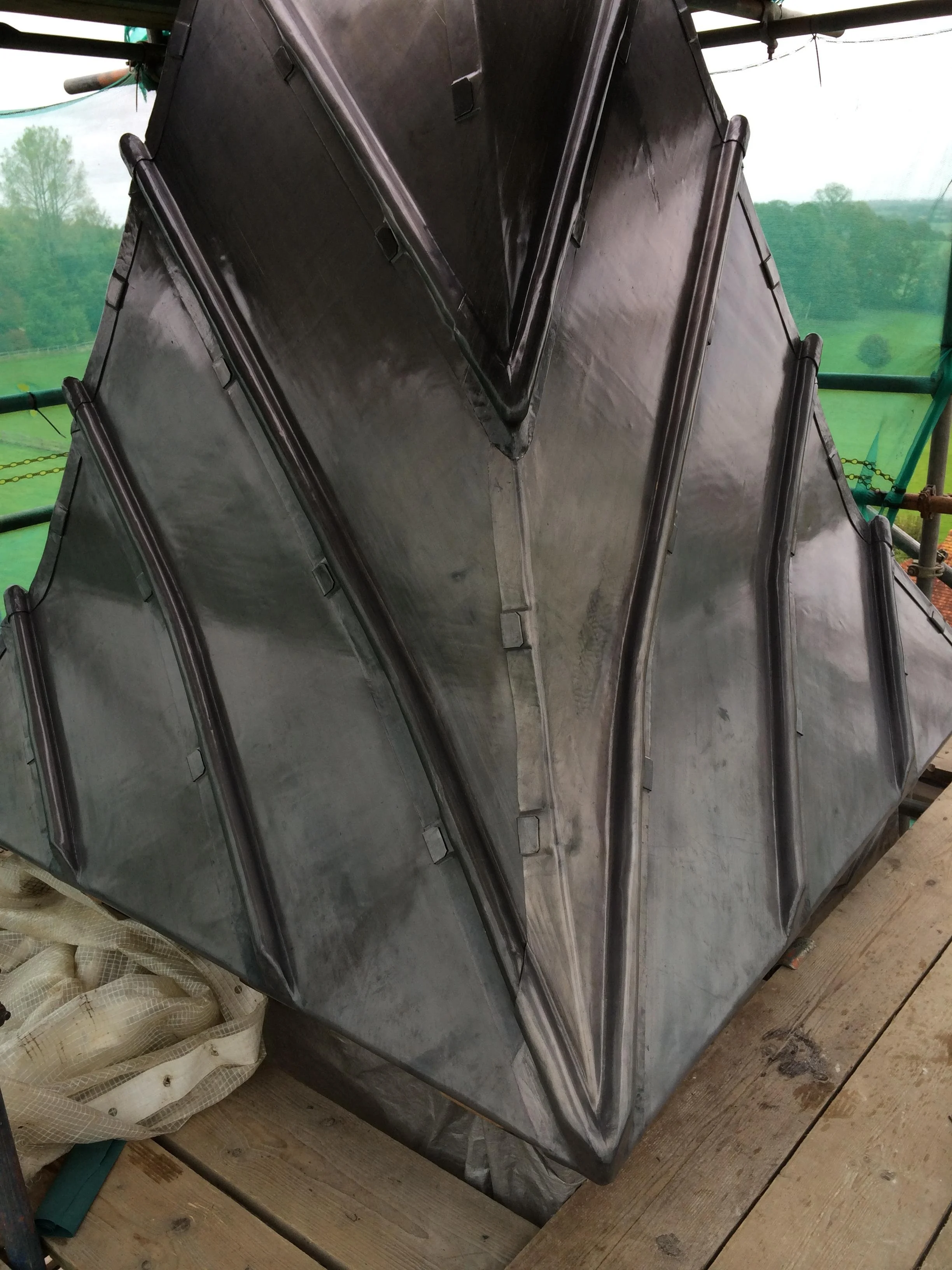 Close-up of a shiny metal geometric structure on a construction site, with scaffolding and a green landscape in the background.