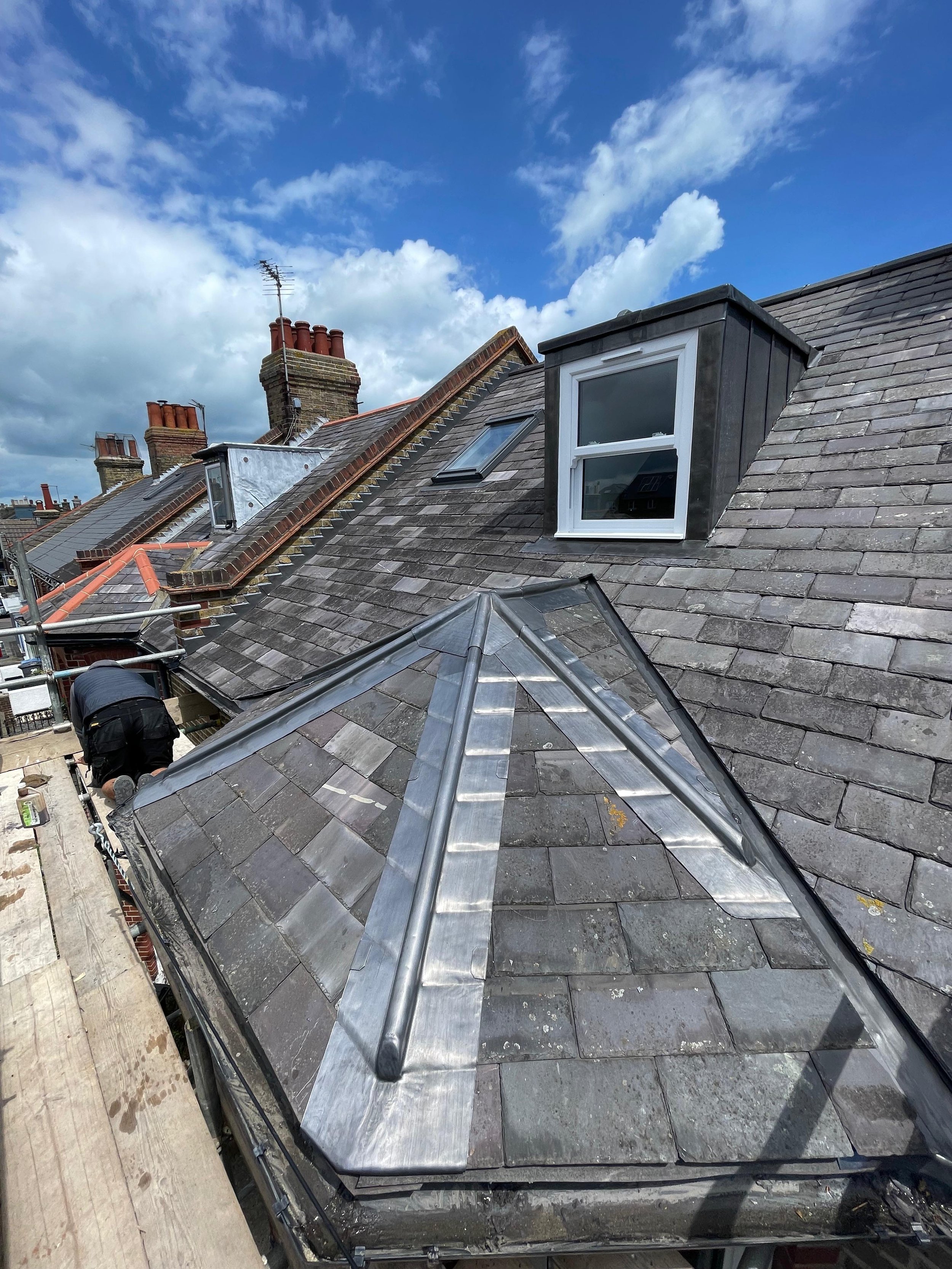 Roof renovation showing slate tiles, a new metal ridge cap, and a worker installing roofing materials under a blue sky with some clouds.