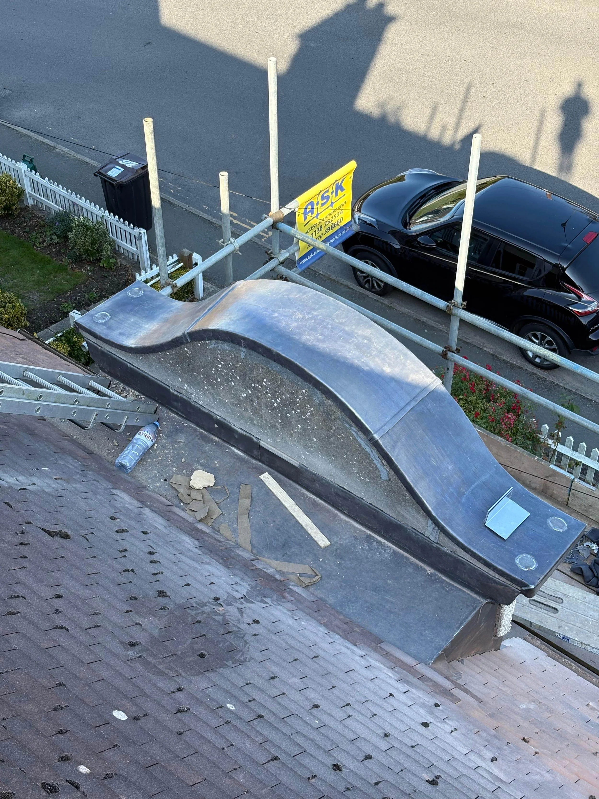 Photo of a rooftop with a skylight, construction materials, and a ladder, taken from above. Below, a street with parked black car and a yellow sign in the distance.