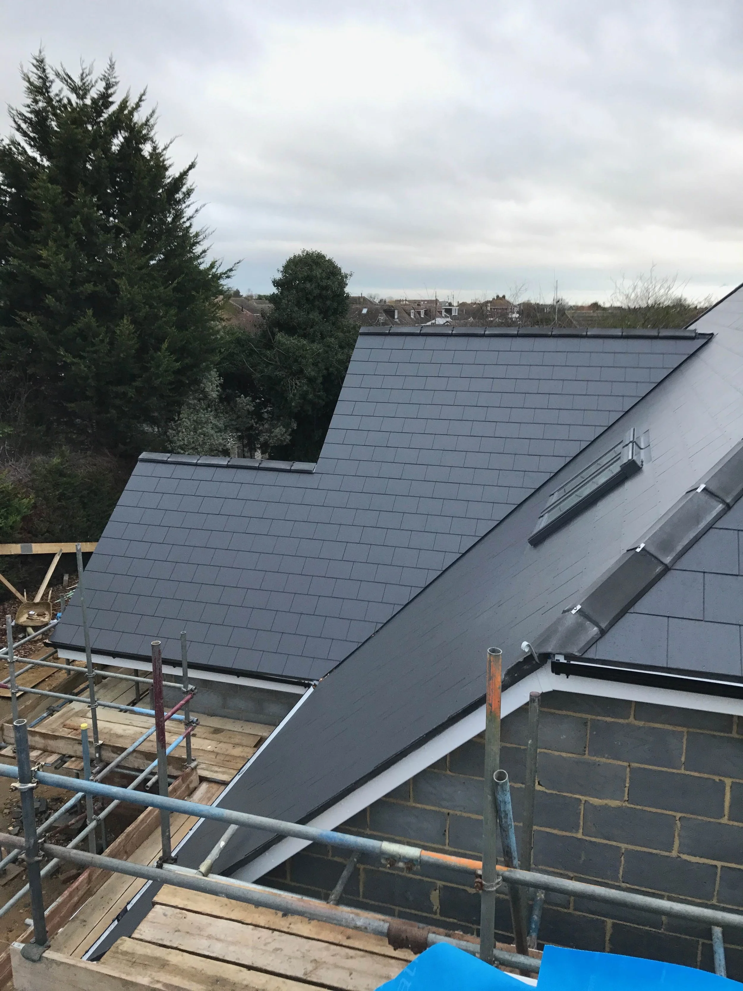 Newly installed dark gray slate roofing on a house with scaffolding, cloudy sky, trees and neighboring houses in the background.