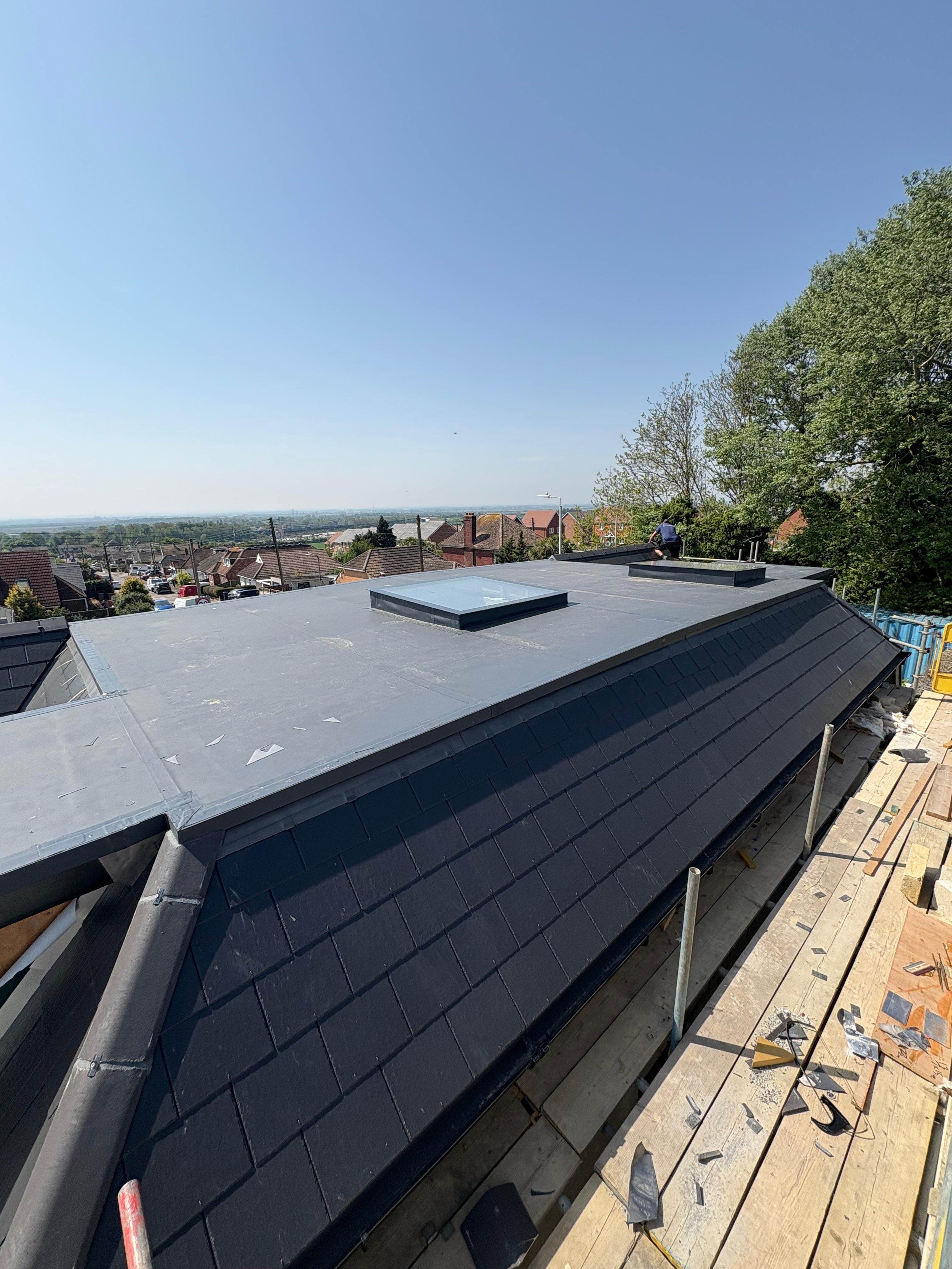 Construction workers installing a new metal roof on a residential building under a clear blue sky, with neighboring houses and trees in the background.