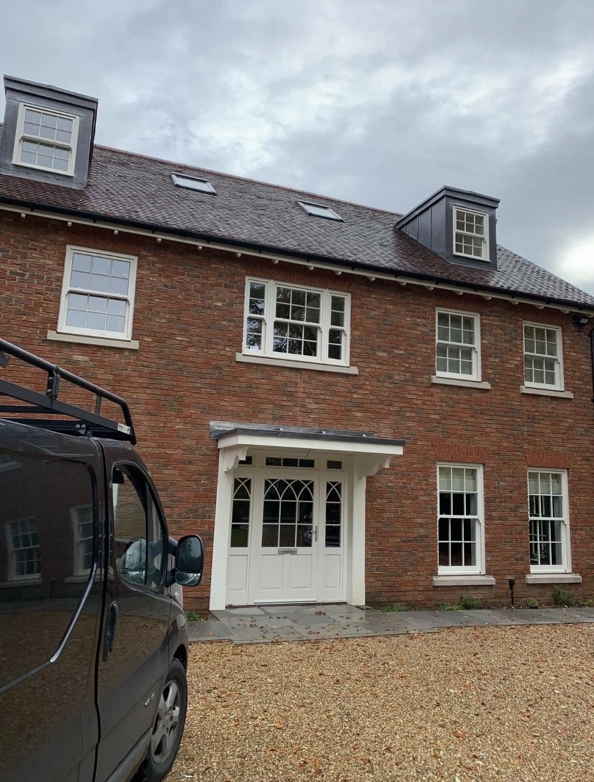 A brick house with white-framed windows and a white door, with a vehicle partially visible on the left side, a gravel driveway, and a cloudy sky above.