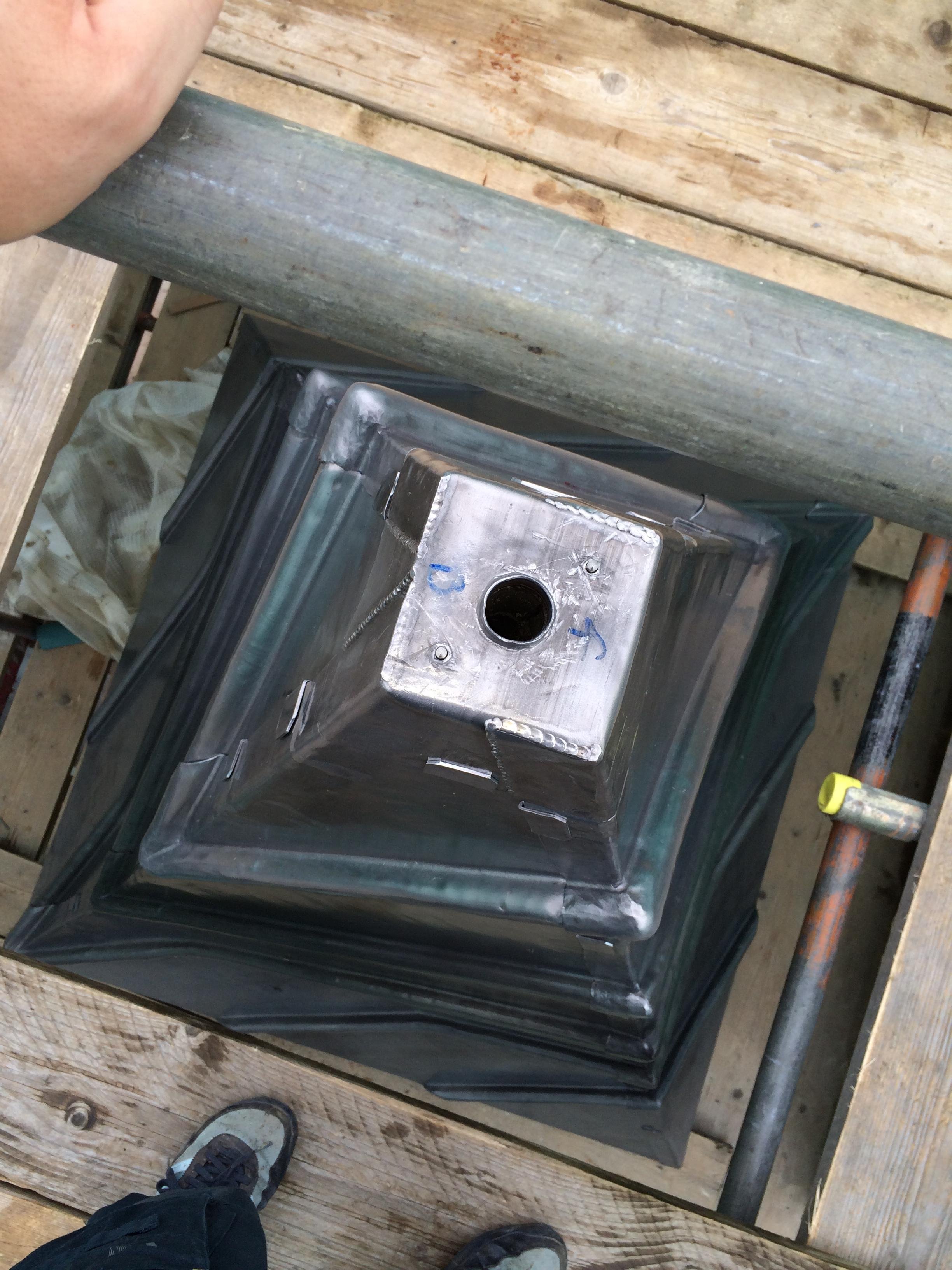 A metal rooftop vent cap with a circular opening in the center, installed on a black metal roof, viewed from above. Scaffolding and wooden planks surround the area.