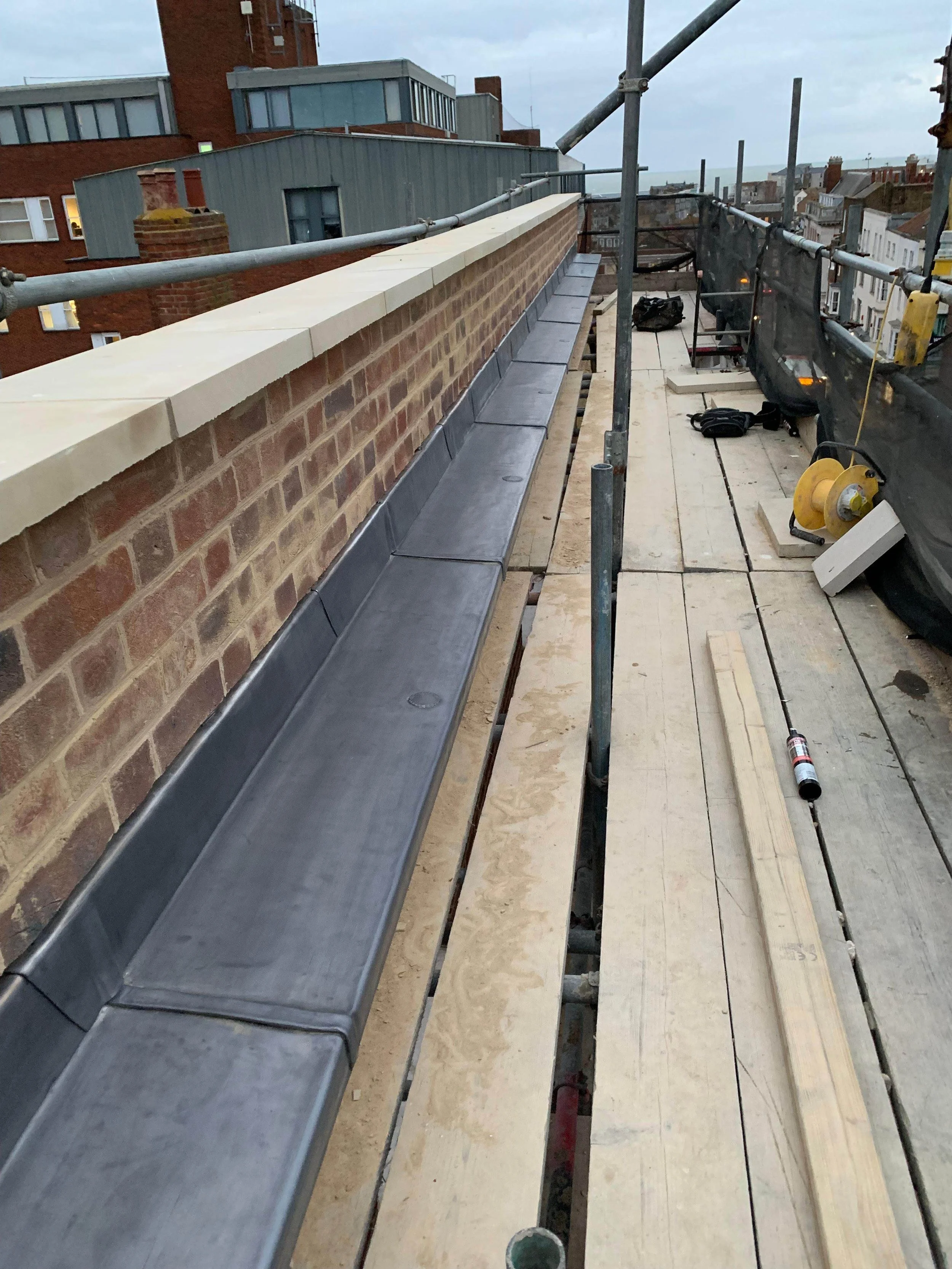 Construction site on a rooftop with scaffolding, tools, and materials, including a long gutter system being installed along the edge of the roof.