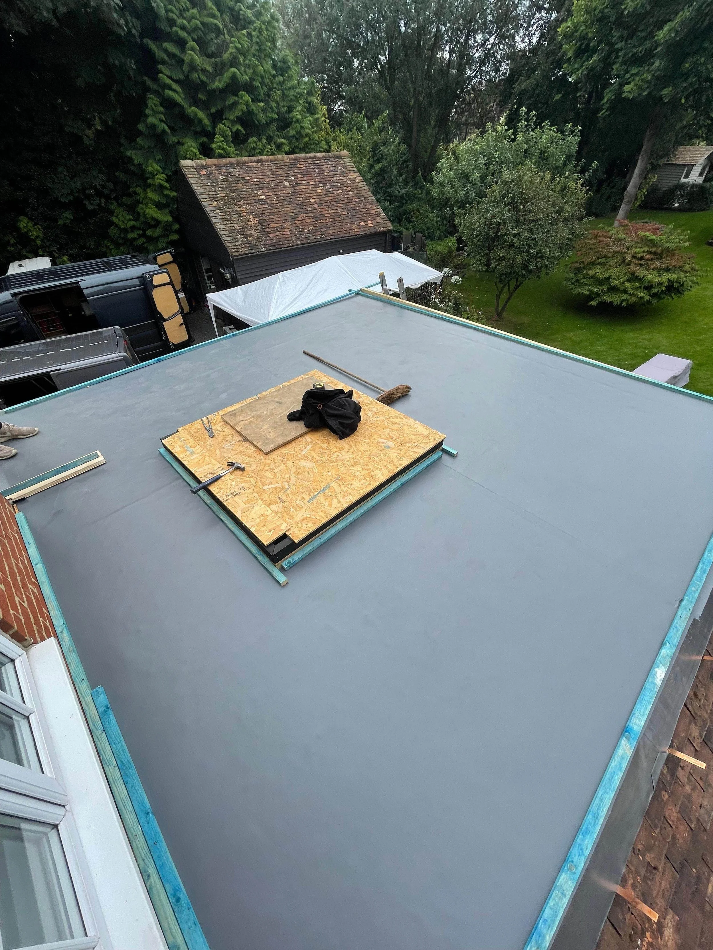 View of a flat rooftop under construction with a plywood hatch, tools, and a broom, overlooking a backyard with trees and a shed.