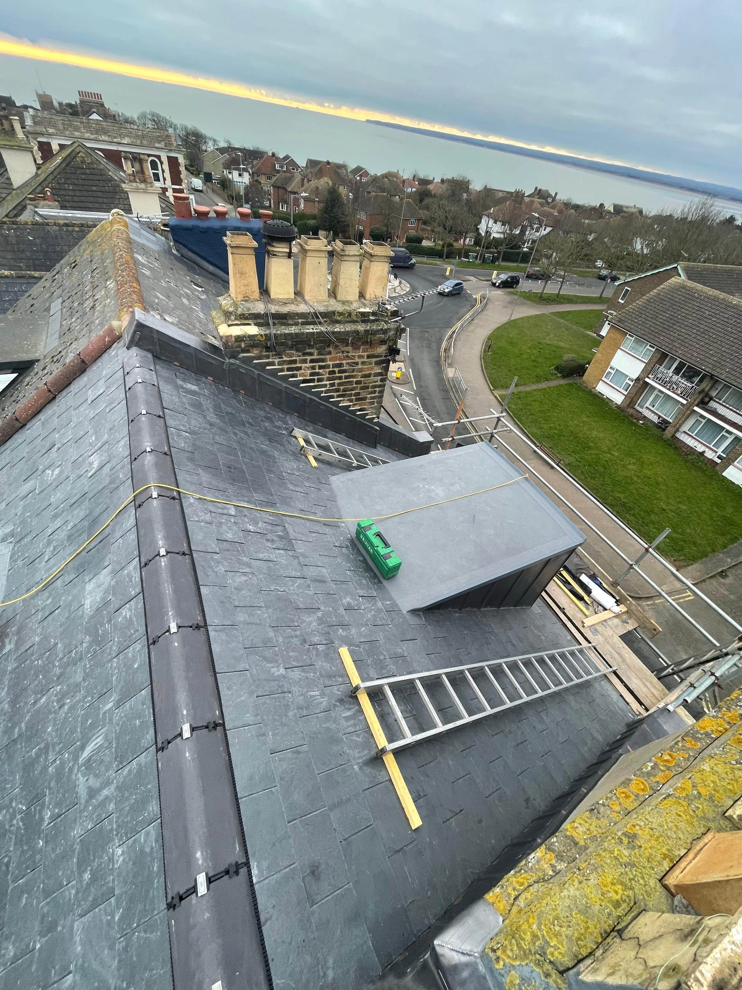 Construction or repair work on a roof with tools and equipment, overlooking a residential neighborhood and a body of water in the background.
