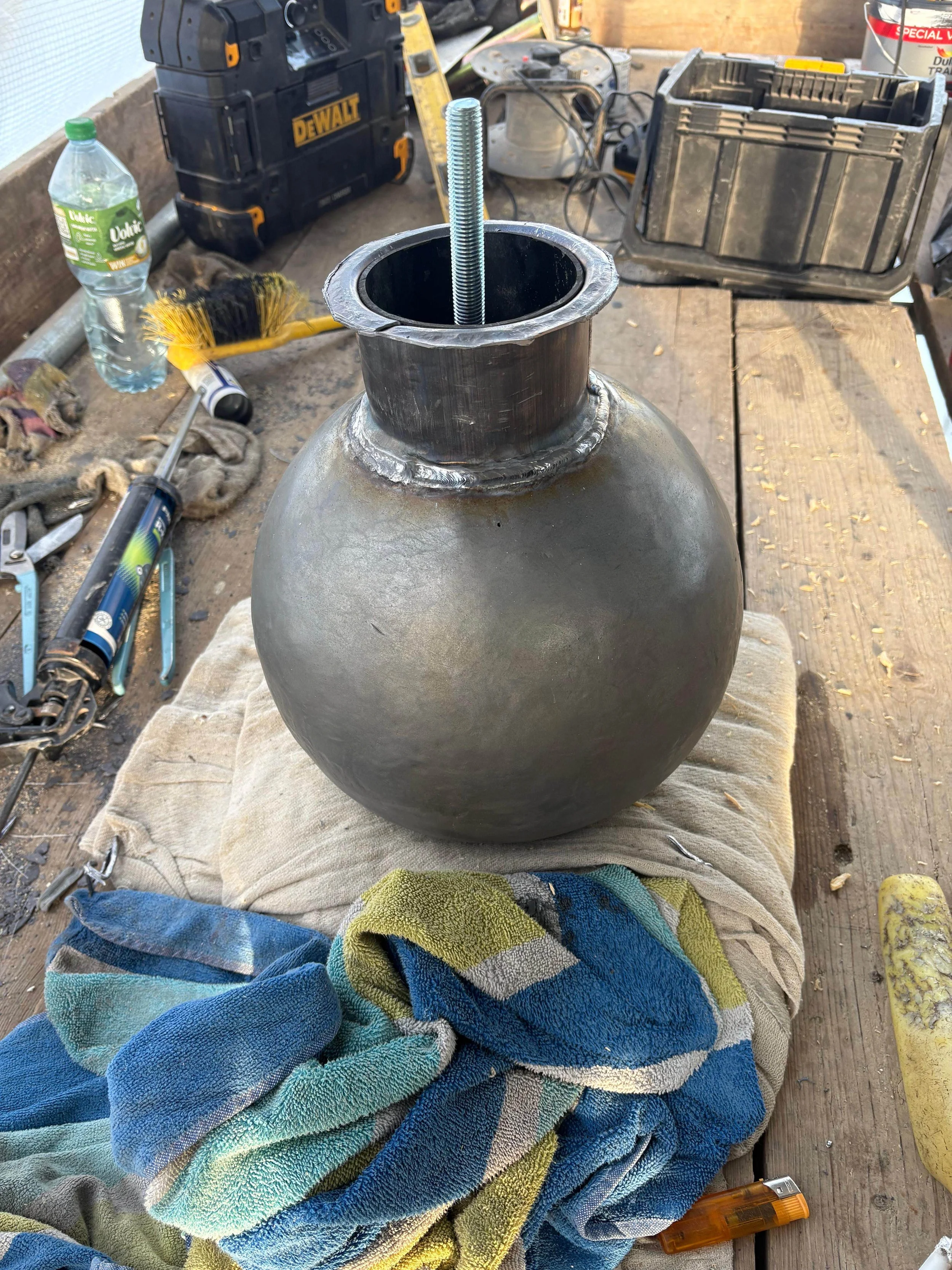 A spherical metal object with an open top and a threaded bolt inside, sitting on a towel on a wooden workbench surrounded by tools and equipment.