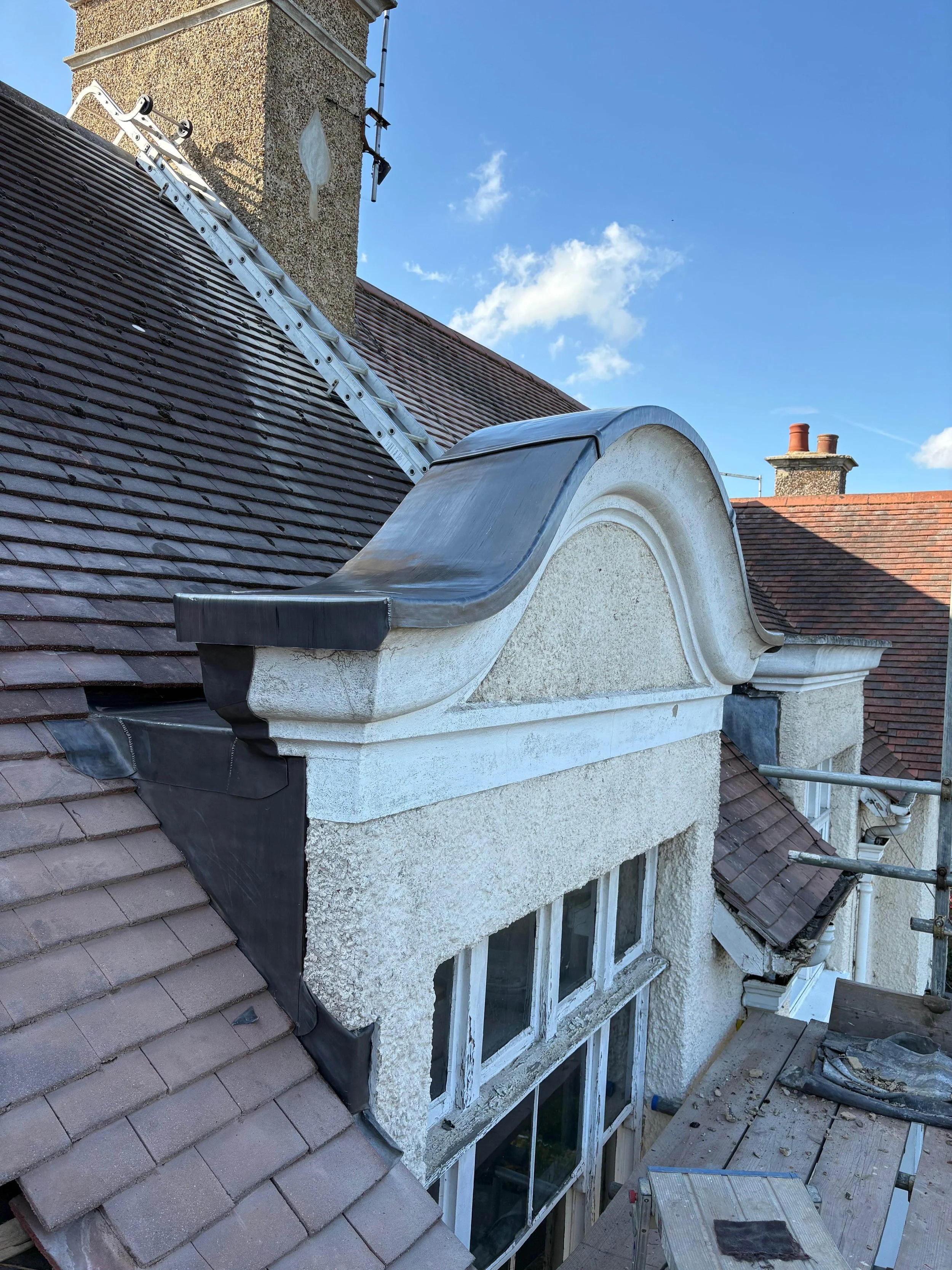 Construction work on a rooftop with scaffolding, showing a decorative window bay with stucco finish and roofing materials.