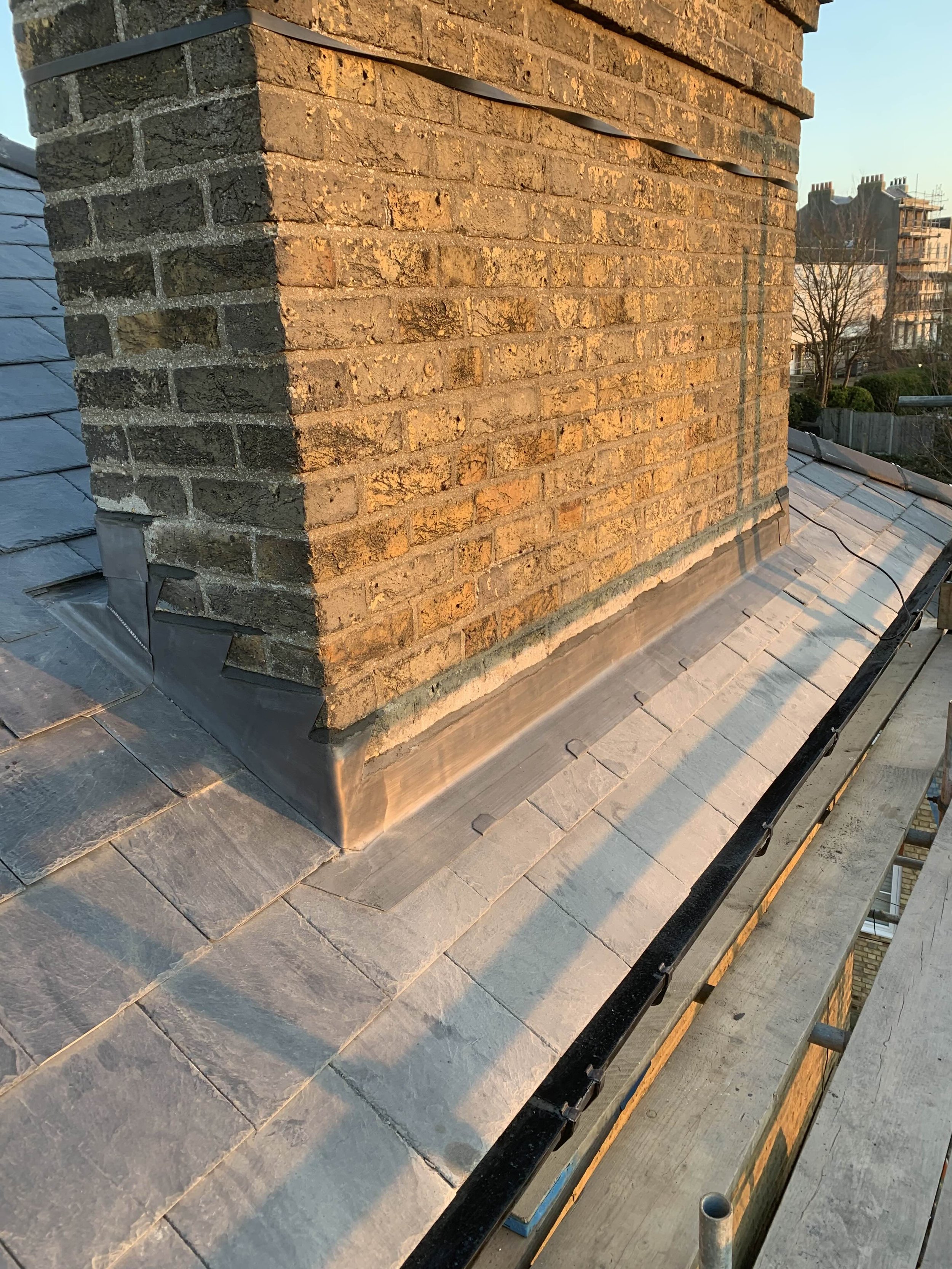 Close-up of a brick chimney on a rooftop with slate tiles, showing the transition from the brick structure to the roof, captured during sunset.