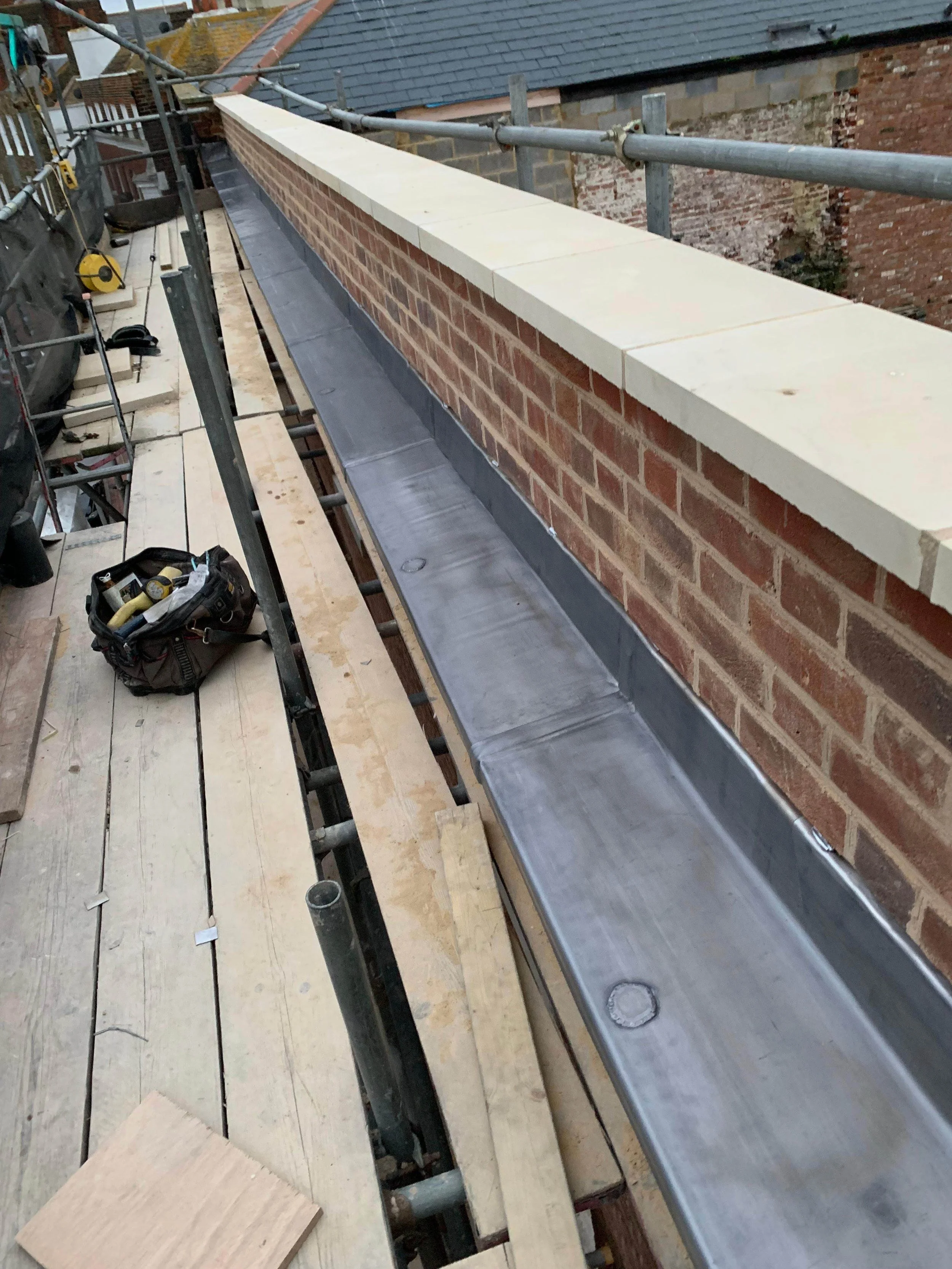 Construction site on a rooftop with scaffolding, showing a brick wall with a newly installed white brick cap and a metal flashing installed at its base; tools and work materials are present on the wooden planks.