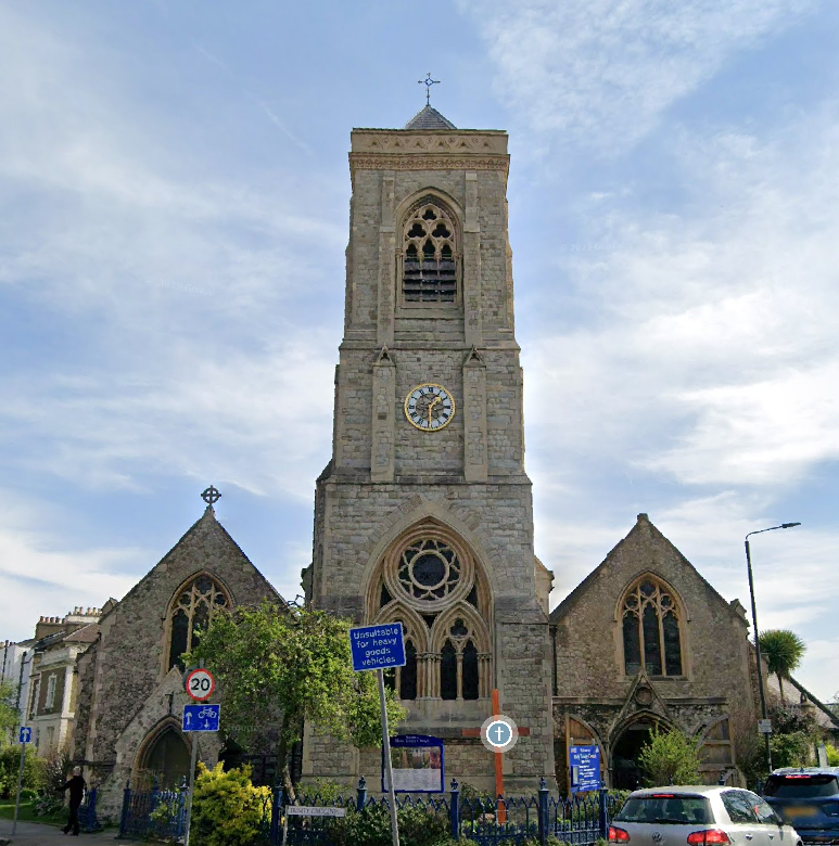 A historic stone church with a tall steeple, clock, and arched windows, situated in a small town street with parked cars and trees.