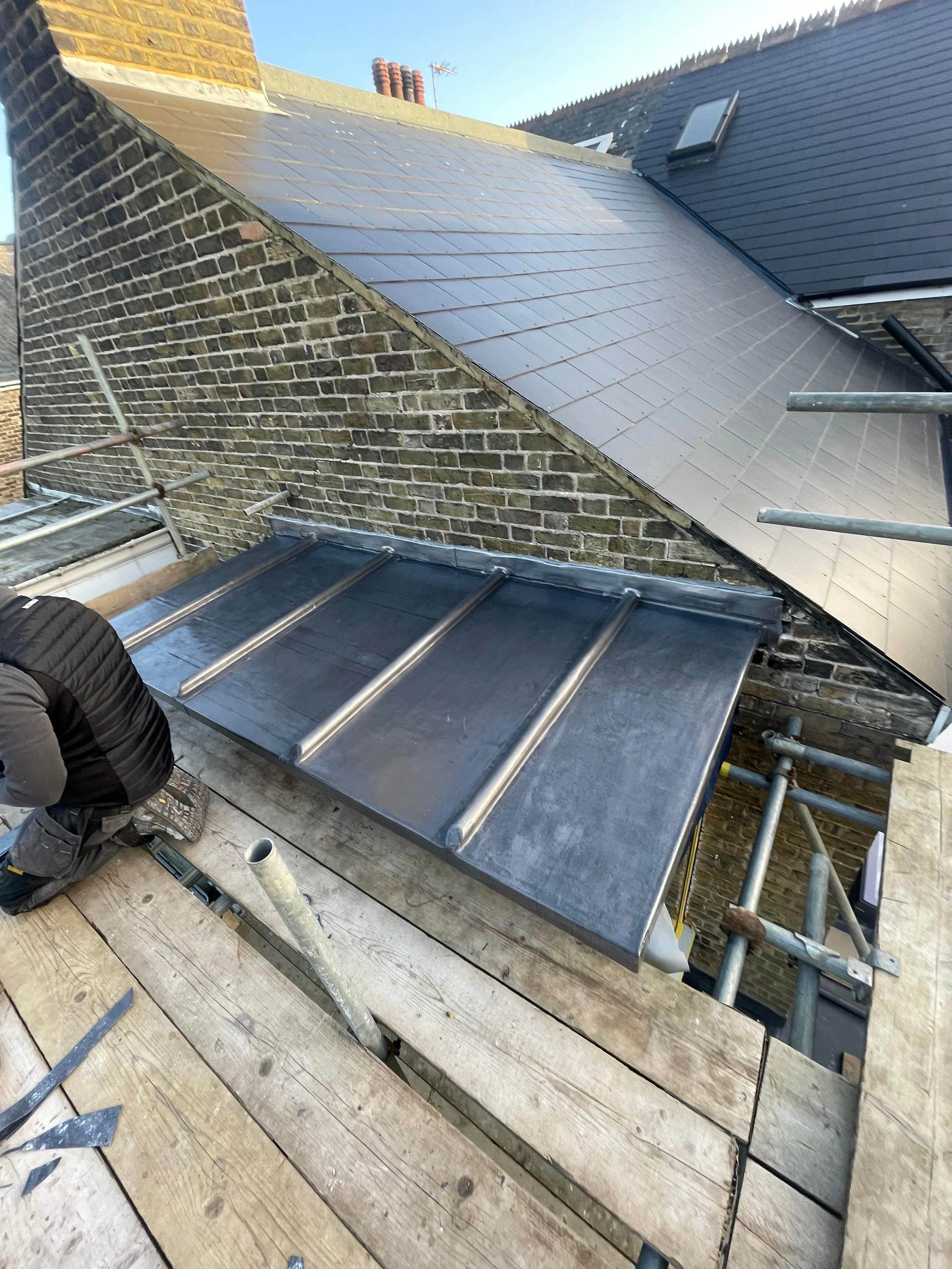 Construction worker installing a metal roof on a building with scaffolding and wooden planks nearby.
