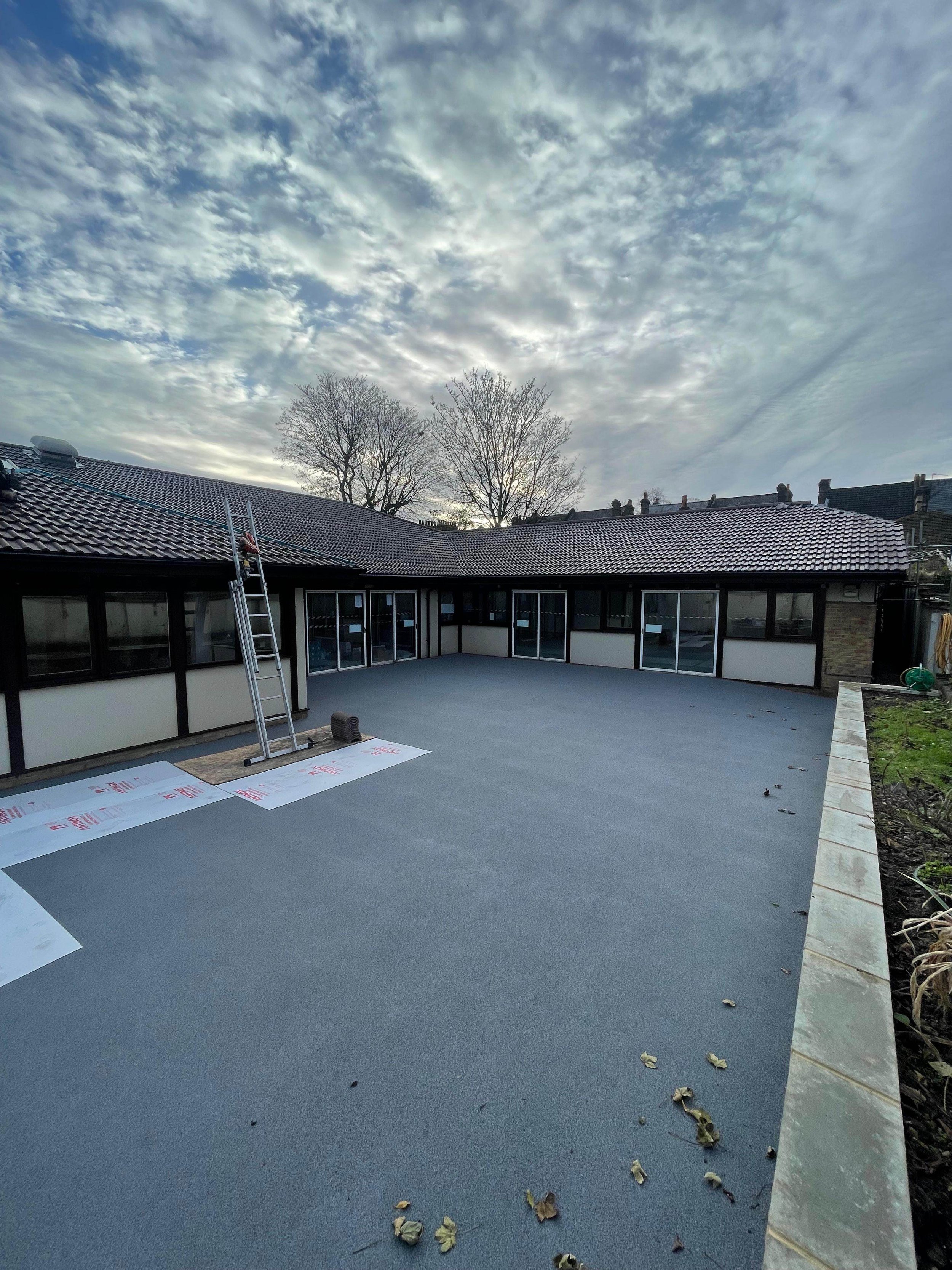 A newly laid flat roof with troweled surface, surrounded by a low wall with a garden bed on the right. A ladder leans against the building, and roofing materials are on the ground. The sky is partly cloudy with trees visible in the background.