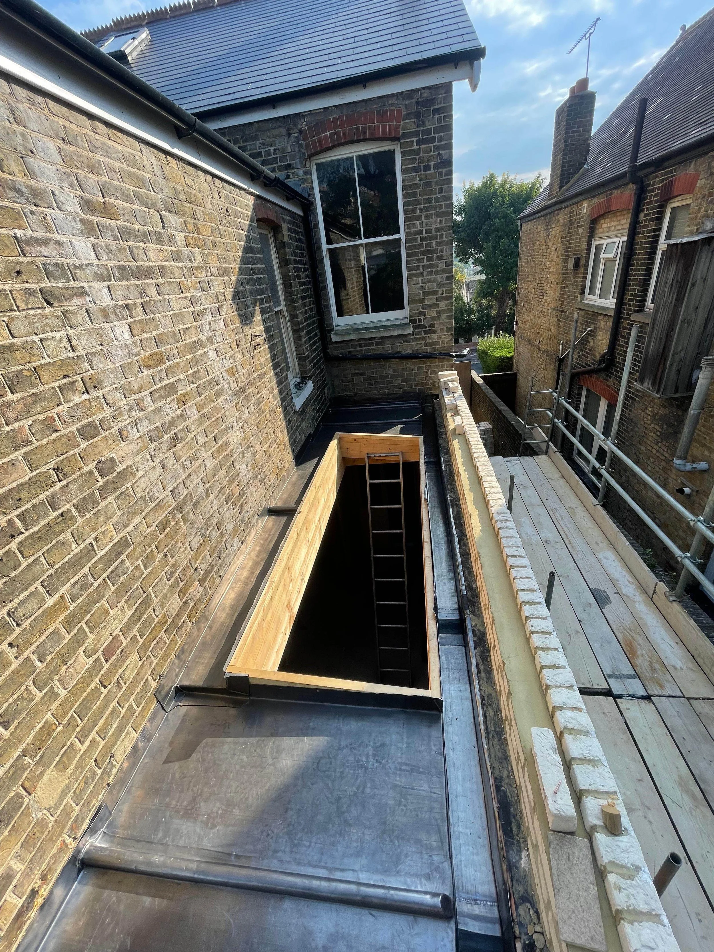 Construction site on a building roof showing a rectangular wooden frame, scaffolding, brick walls, and a window, with a ladder inside the frame.