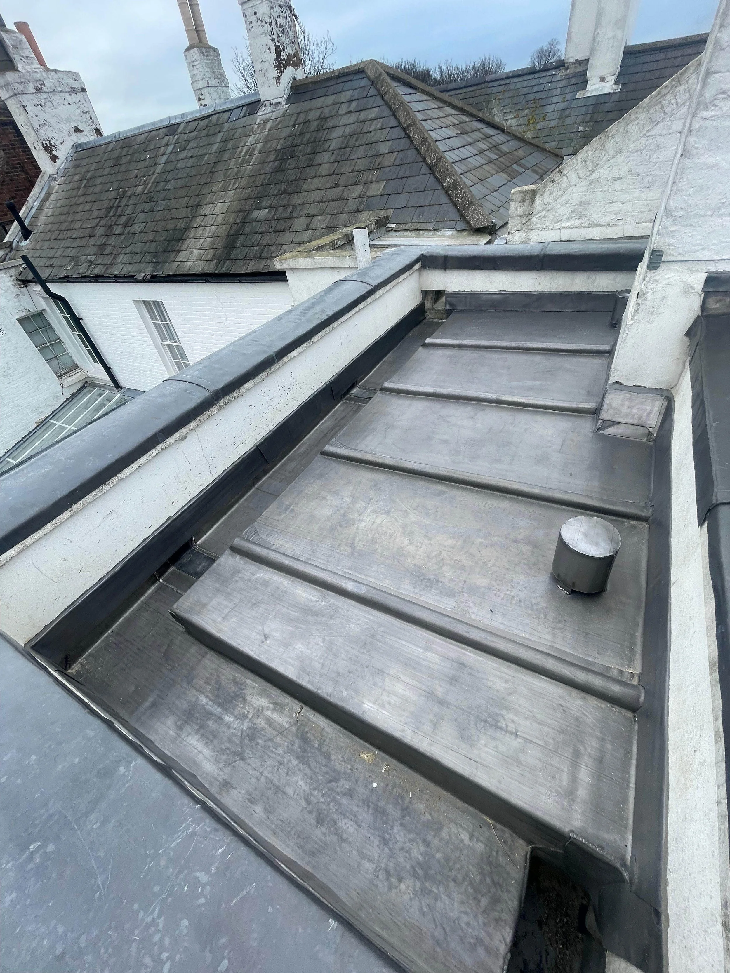 View of a metal flat roof on a building with a vent pipe, with neighboring houses and cloudy sky in background.