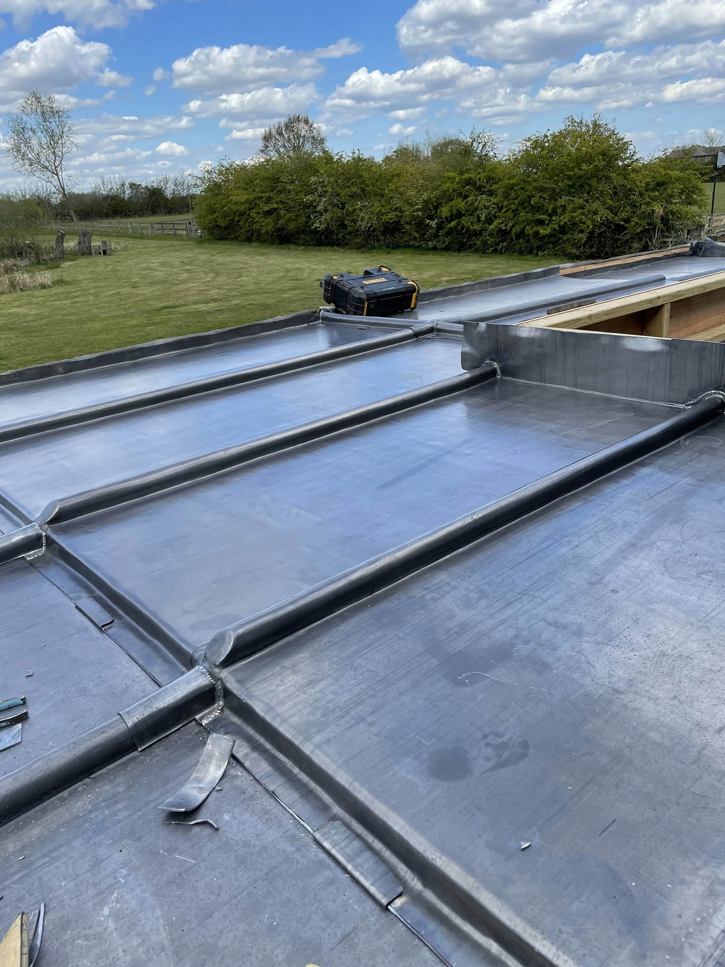 A roof with a black toolbox and metal flashing during construction, with trees and a grassy field in the background under a partly cloudy sky.
