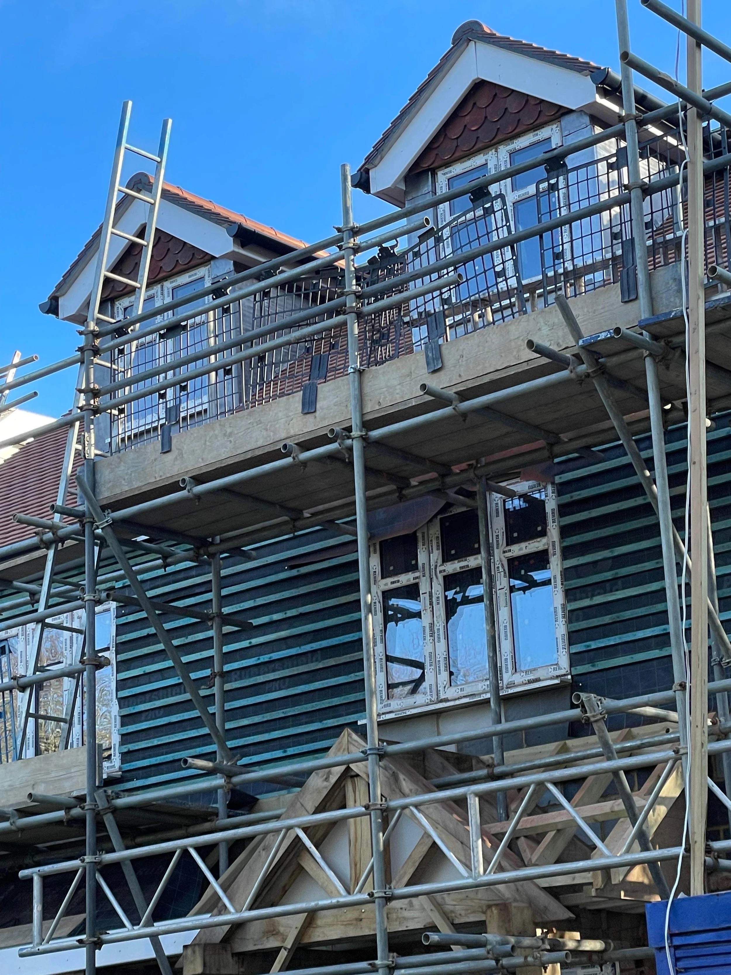 A house under construction with scaffolding around it, showing incomplete exterior walls with black siding, and windows installed on the upper floor. The roof has brown tiles, and part of the structure is covered in blue sky.