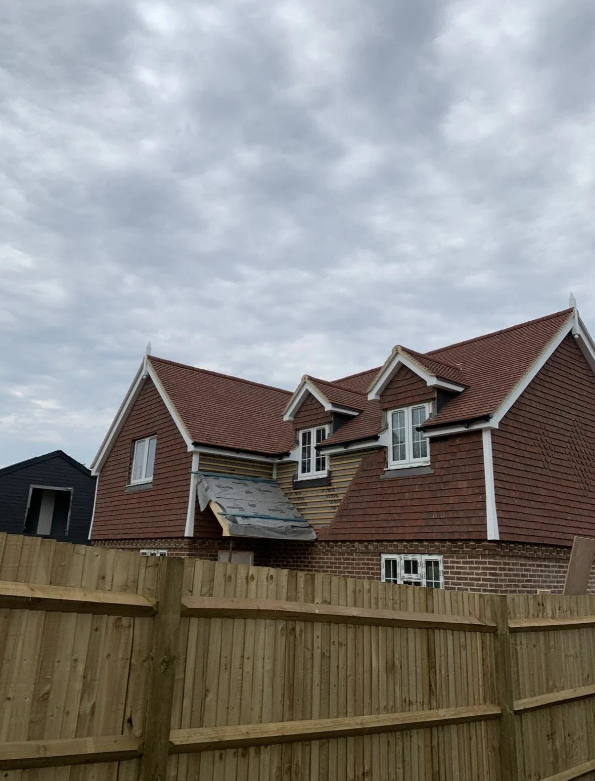 A house under construction with red roof tiles, white window frames, and a partially completed exterior wall. A wooden fence surrounds the property, and the sky is cloudy.
