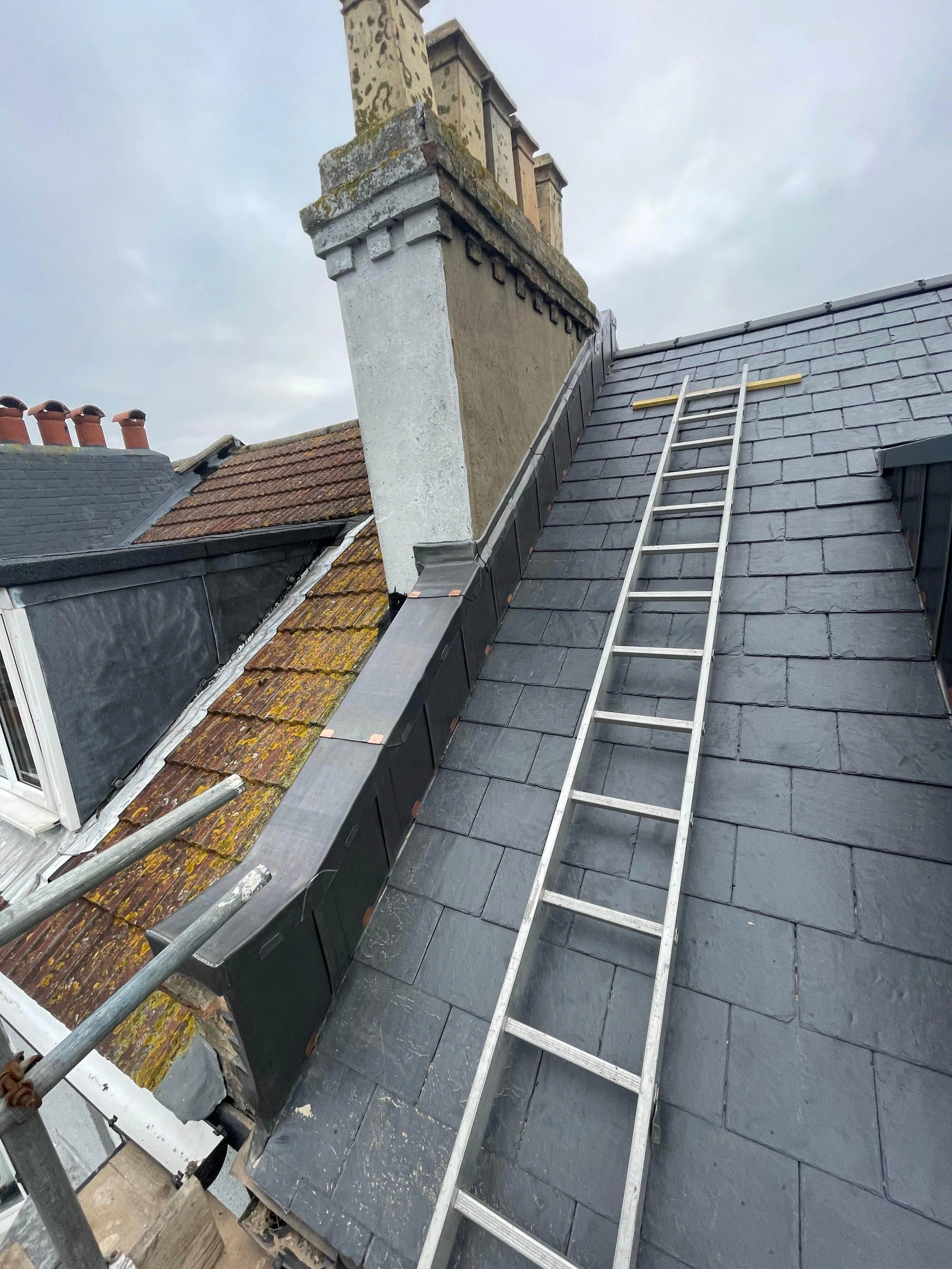 View of a roof with slate tiles, a chimney, utility ladders, and scaffolding, on an overcast day.