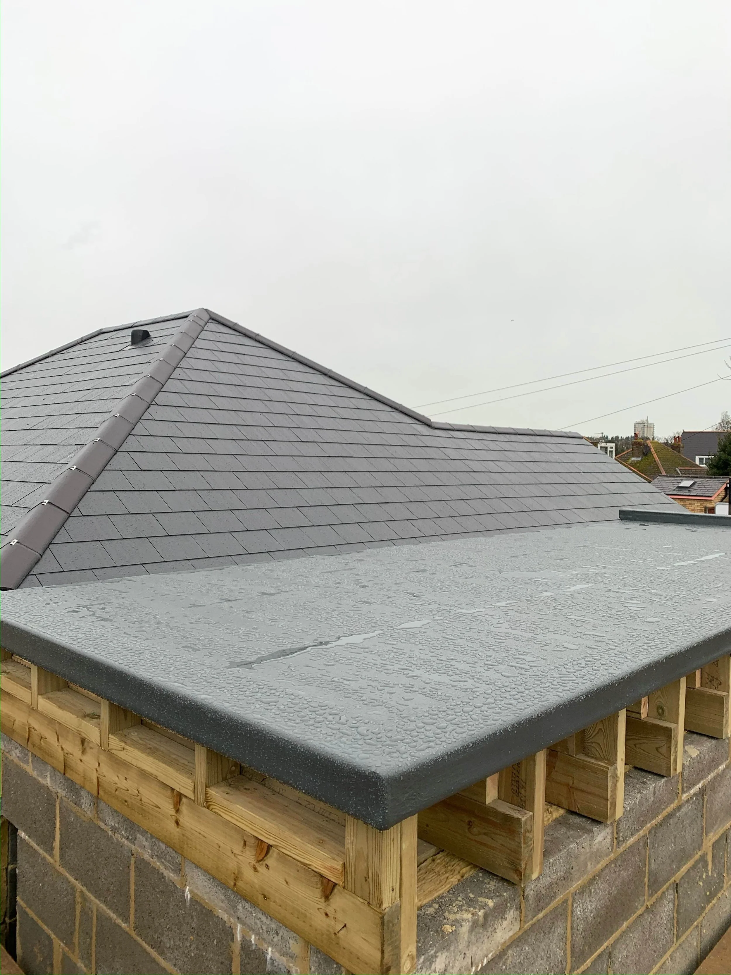Close-up of a house roof with dark gray, new shingles and a flat chimney cap on the edge, with wooden framing and a brick base beneath. Overcast sky in the background.